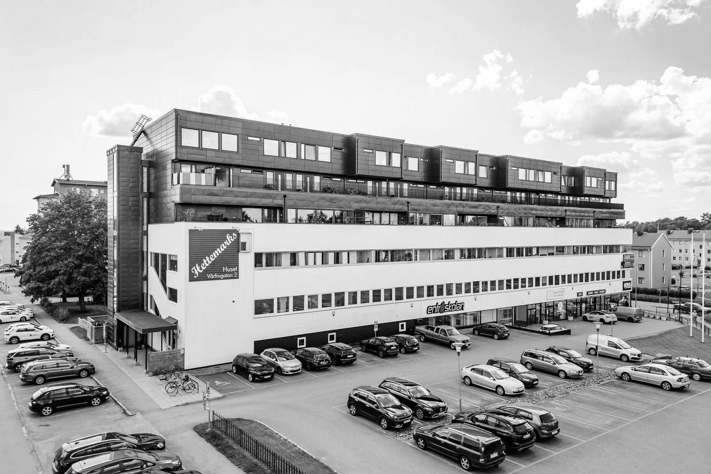 Black-and-white photo of a modern, multi-story building with commercial space on the ground floor and residential or office units above. The building features large windows and a parking lot with several cars and bicycles in front.
