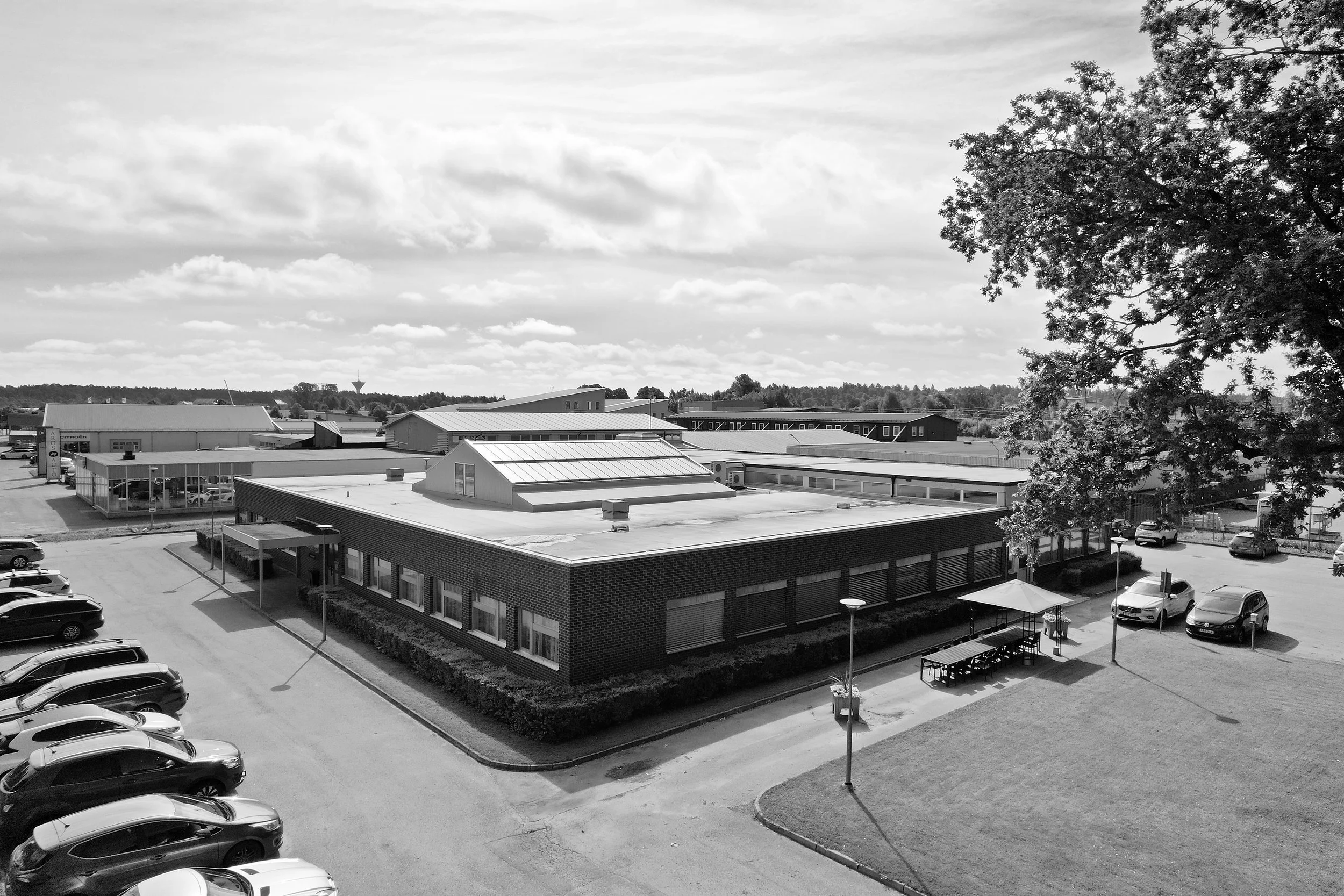 Black and white aerial view of an industrial or commercial complex with parked cars, a small outdoor seating area with tables, and large buildings with flat roofs, some with skylights, surrounded by trees and a cloudy sky.