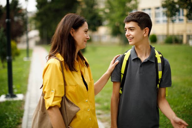A woman and a teenage boy talking outdoors on a pathway with green grass and trees, she is wearing a yellow jacket, he is wearing a gray shirt with a yellow backpack.