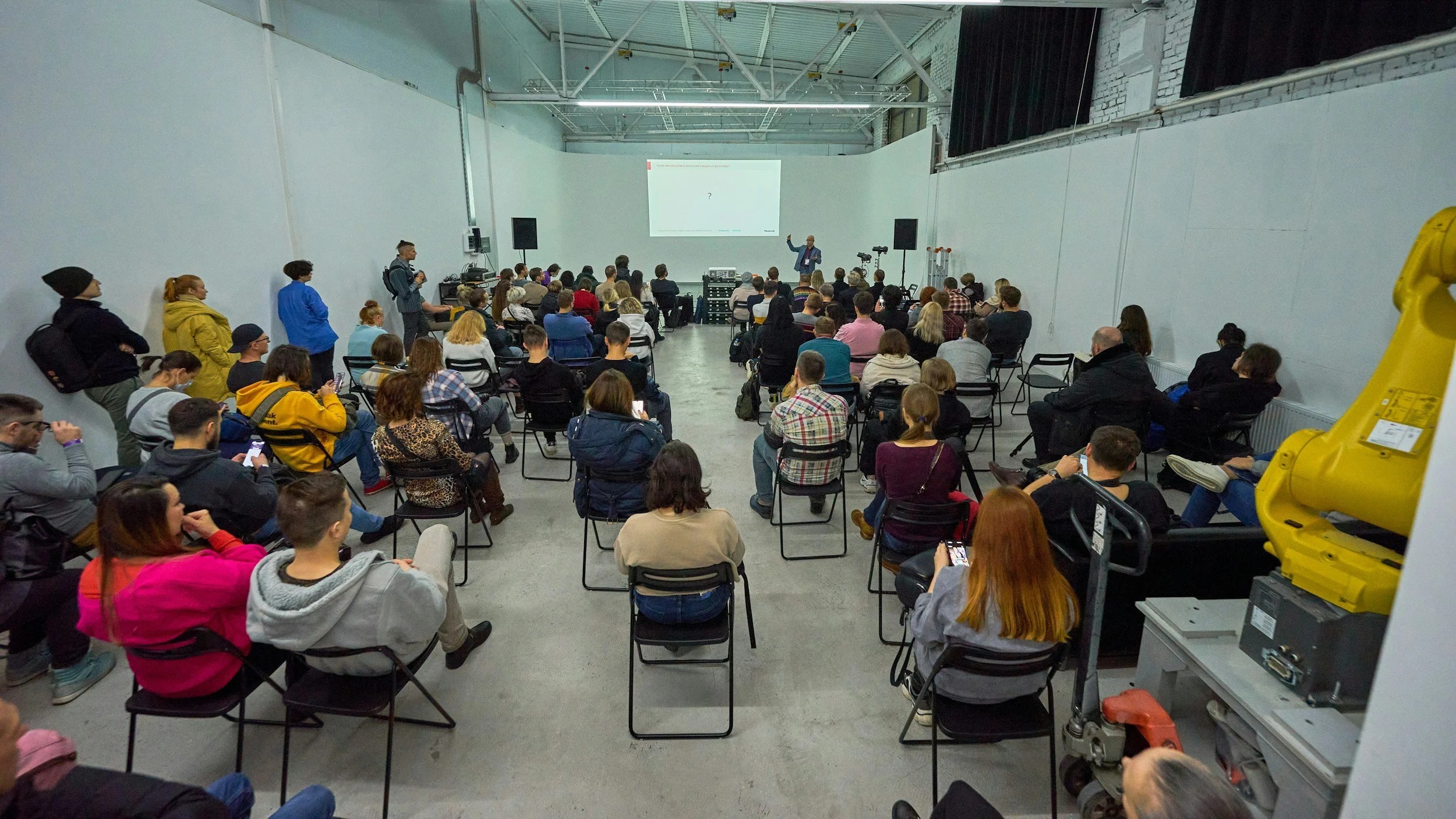 A large group of people attending a presentation in a spacious industrial-style room with white walls and high ceiling, seated on black chairs facing a speaker at the front, with some standing along the walls. The speaker is gesturing near a projector screen displaying a slide, with cameras and audio equipment set up nearby.
