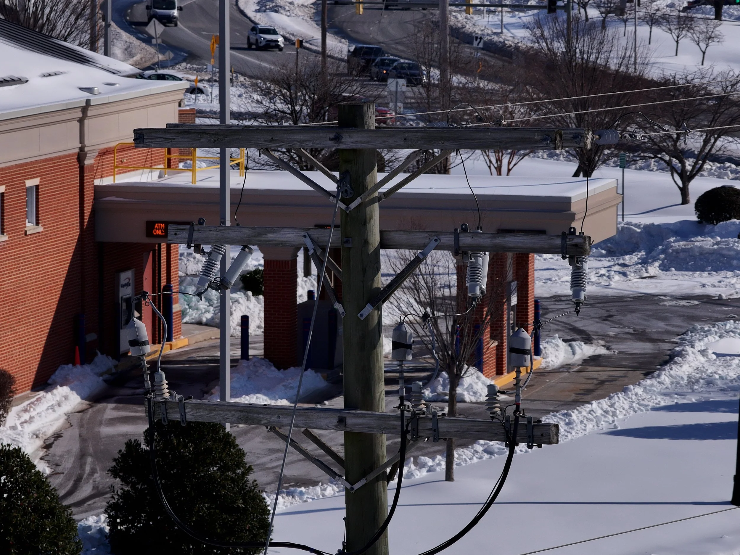 Electric power lines and transformers mounted on a wooden utility pole in a snowy area near a brick building and parking lot.