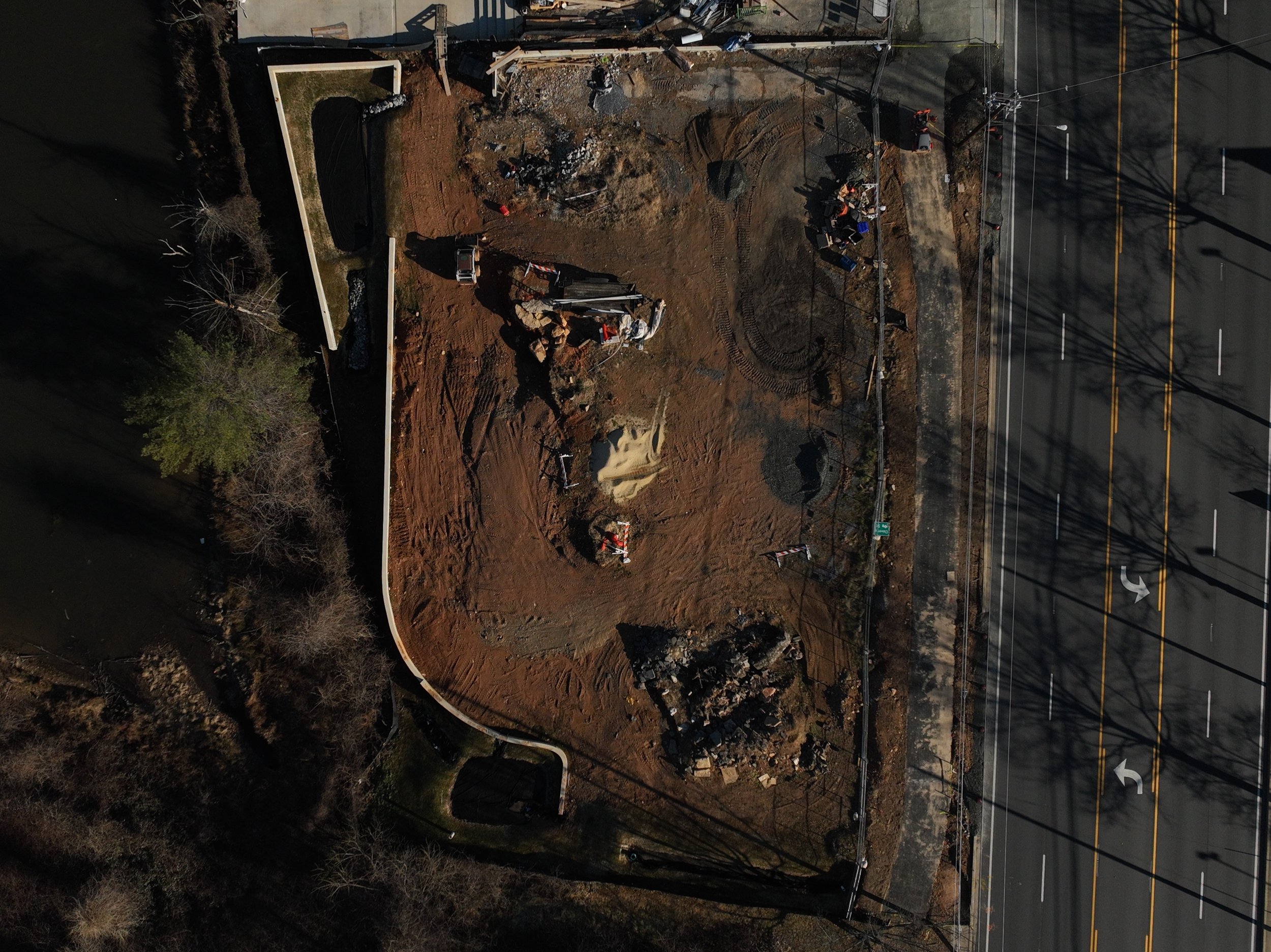An aerial view of a construction site next to a road, showing excavated earth, construction equipment, and partially built retaining wall.