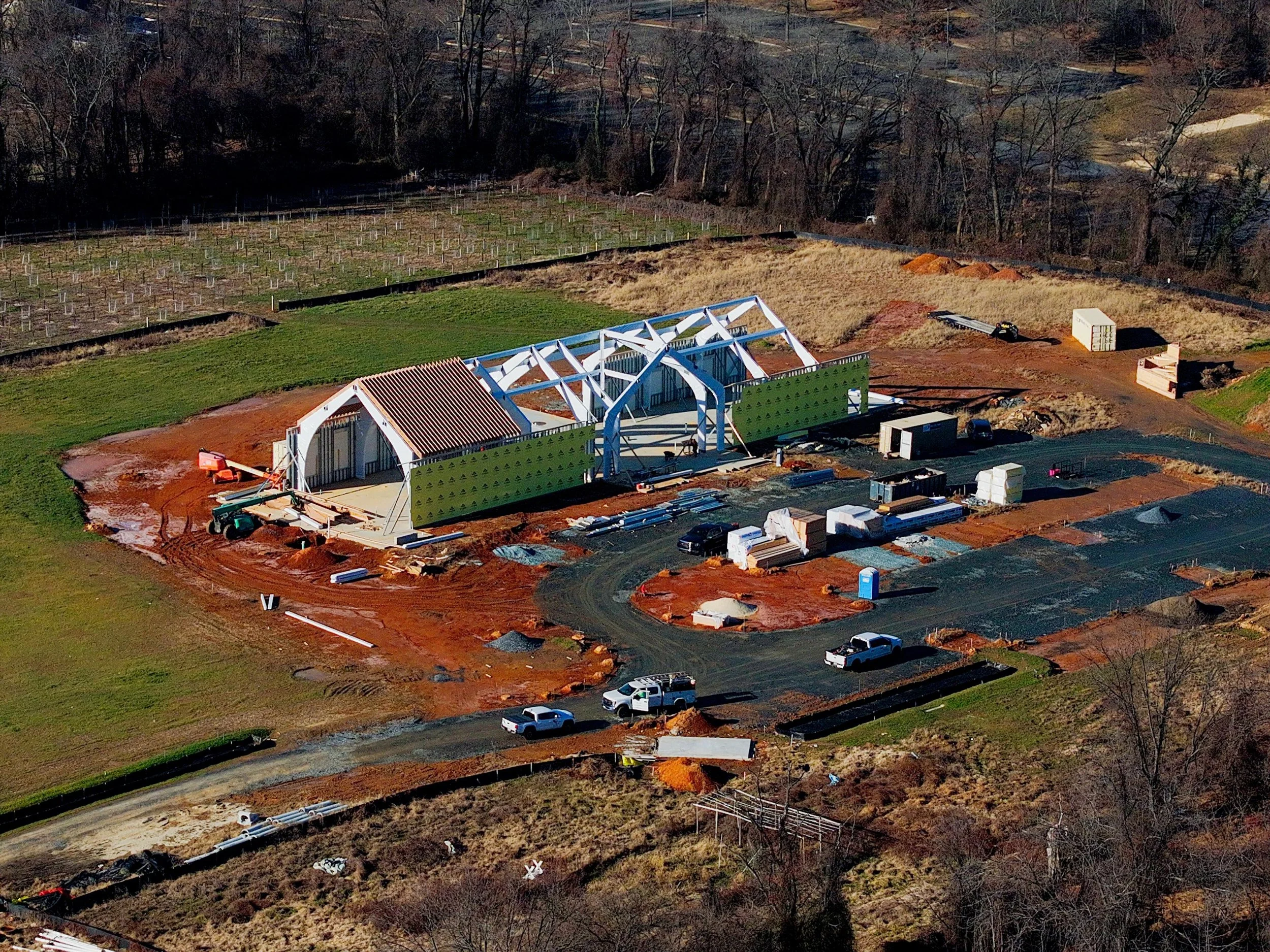 An aerial view of a construction site with a partially built structure featuring white beams and green walls, surrounded by parked vehicles, construction materials, and dirt pathways, with trees and open land in the background.