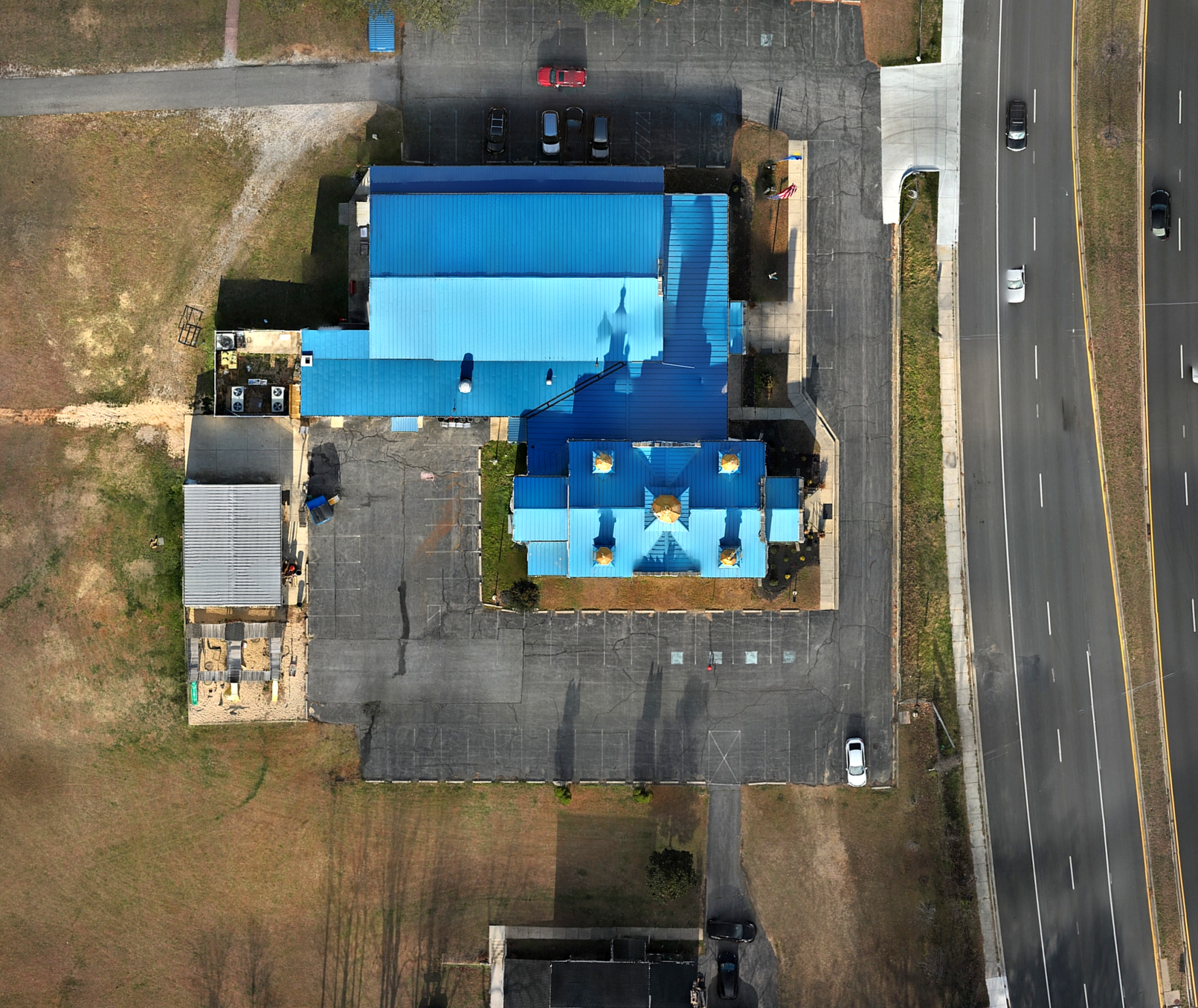 An aerial view of a church building with a blue roof, surrounded by parking lot, grass, and a road with cars.
