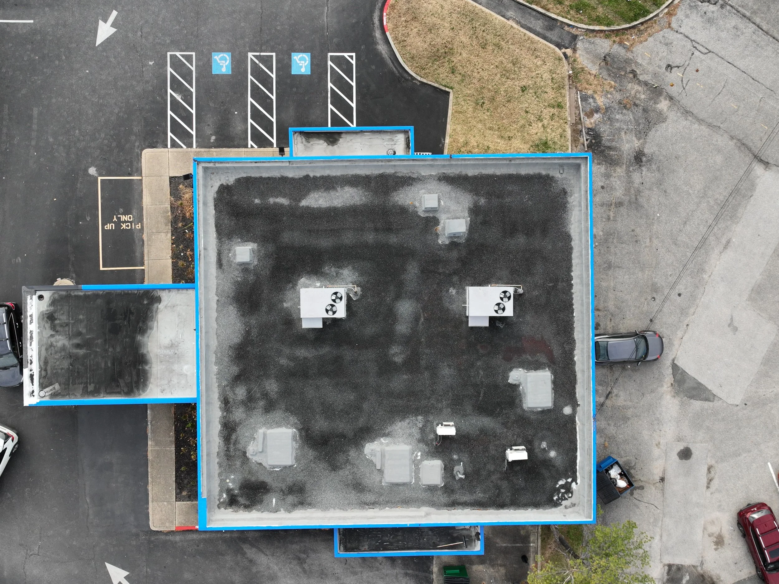 An aerial view of a building with a flat roof, surrounded by a parking lot with marked parking spaces, including designated handicapped spots, and some parked cars.