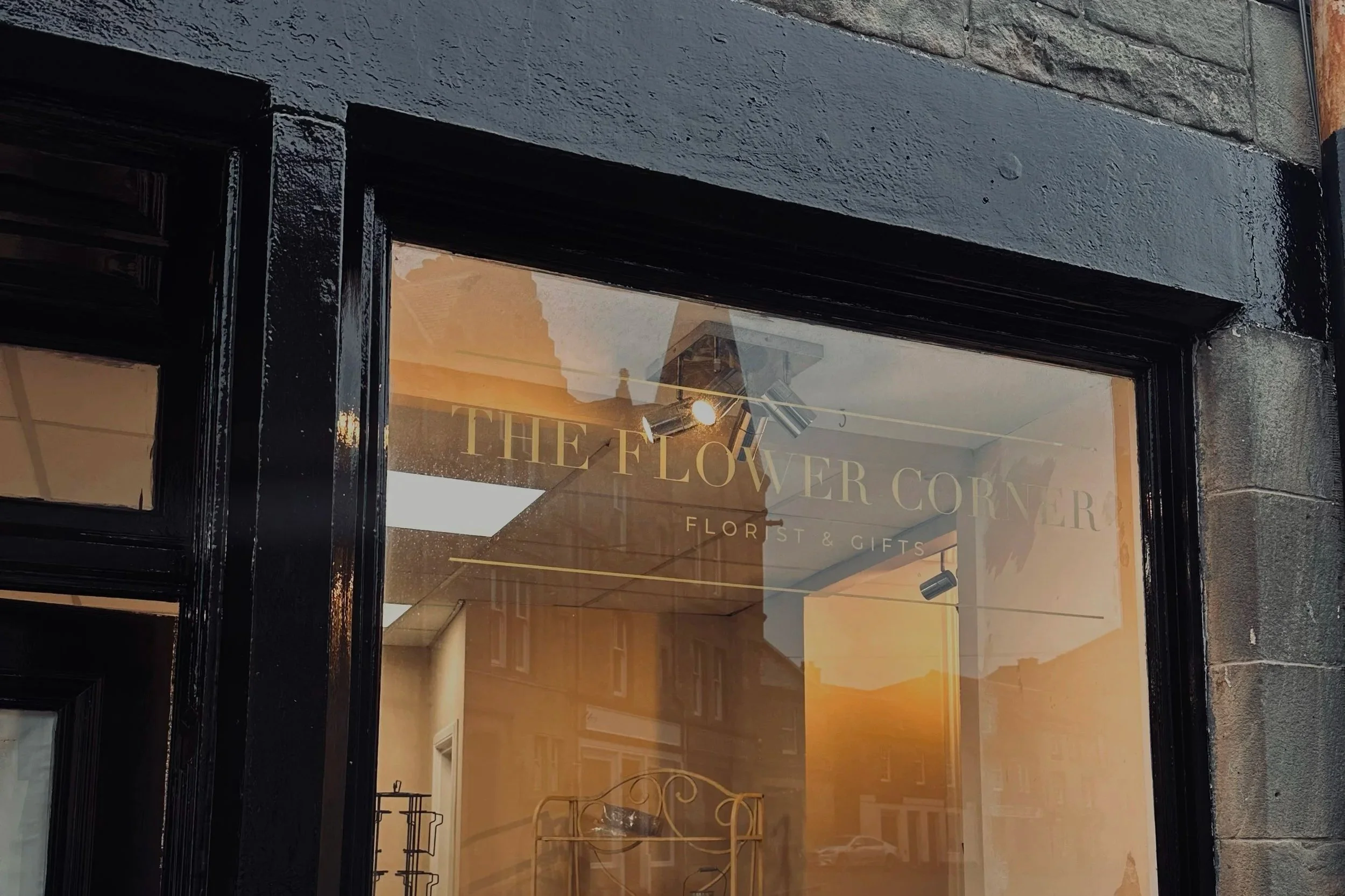 The storefront of a flower shop called "The Flower Corner" with large glass window reflecting a building and sky outside, and interior showing a metal rack.