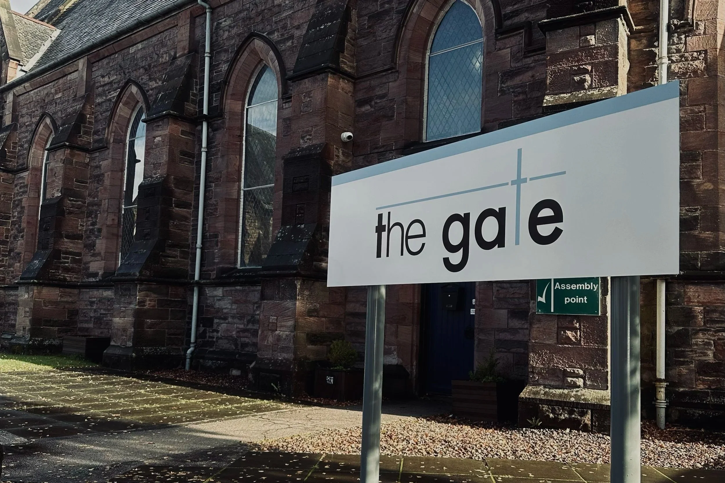 Sign with the words 'the gate' in front of a brick building with arched windows and a small plant in a planter near the door.