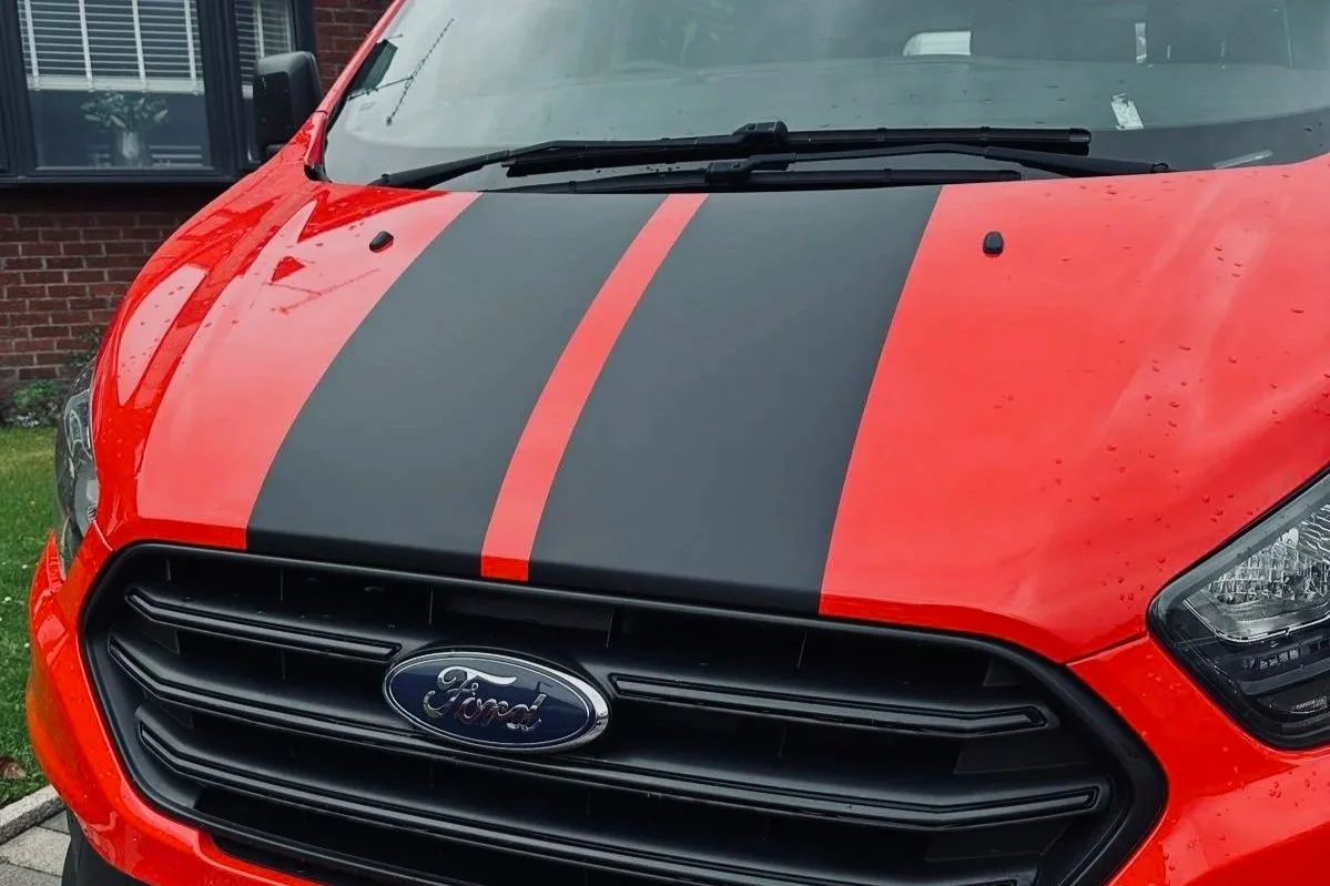 Red Ford vehicle with black racing stripes on the hood, parked outside a brick building.