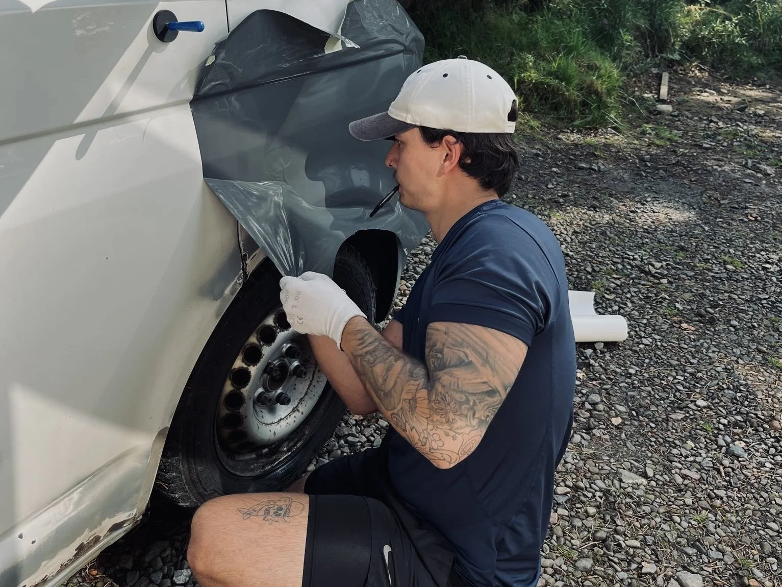 A man with tattoos and a white cap applying a vinyl wrap to a white car's front fender outdoors on rocky ground, with greenery in the background.