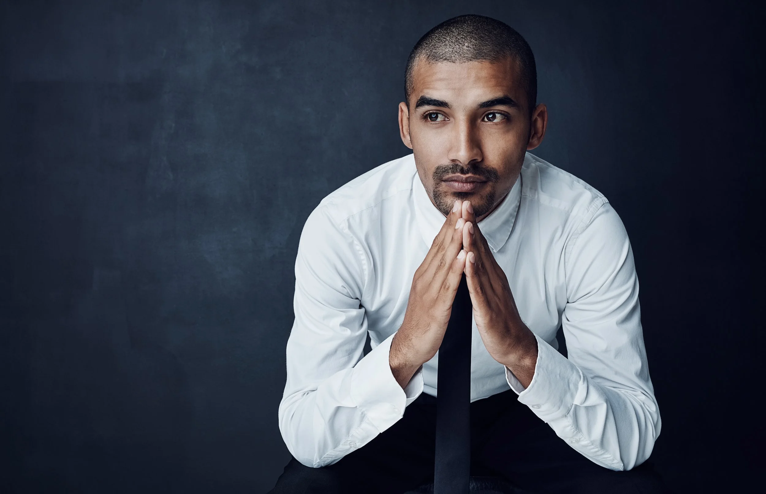 A young man with a shaved head and goatee in a white shirt and black tie, with his hands clasped in front of his face, looking thoughtful against a dark background.