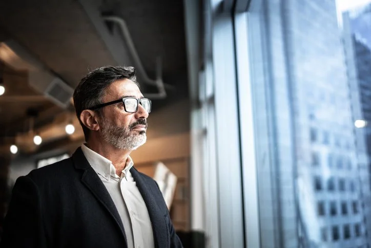 A middle-aged man with glasses and a beard looking out a window in a modern office building.