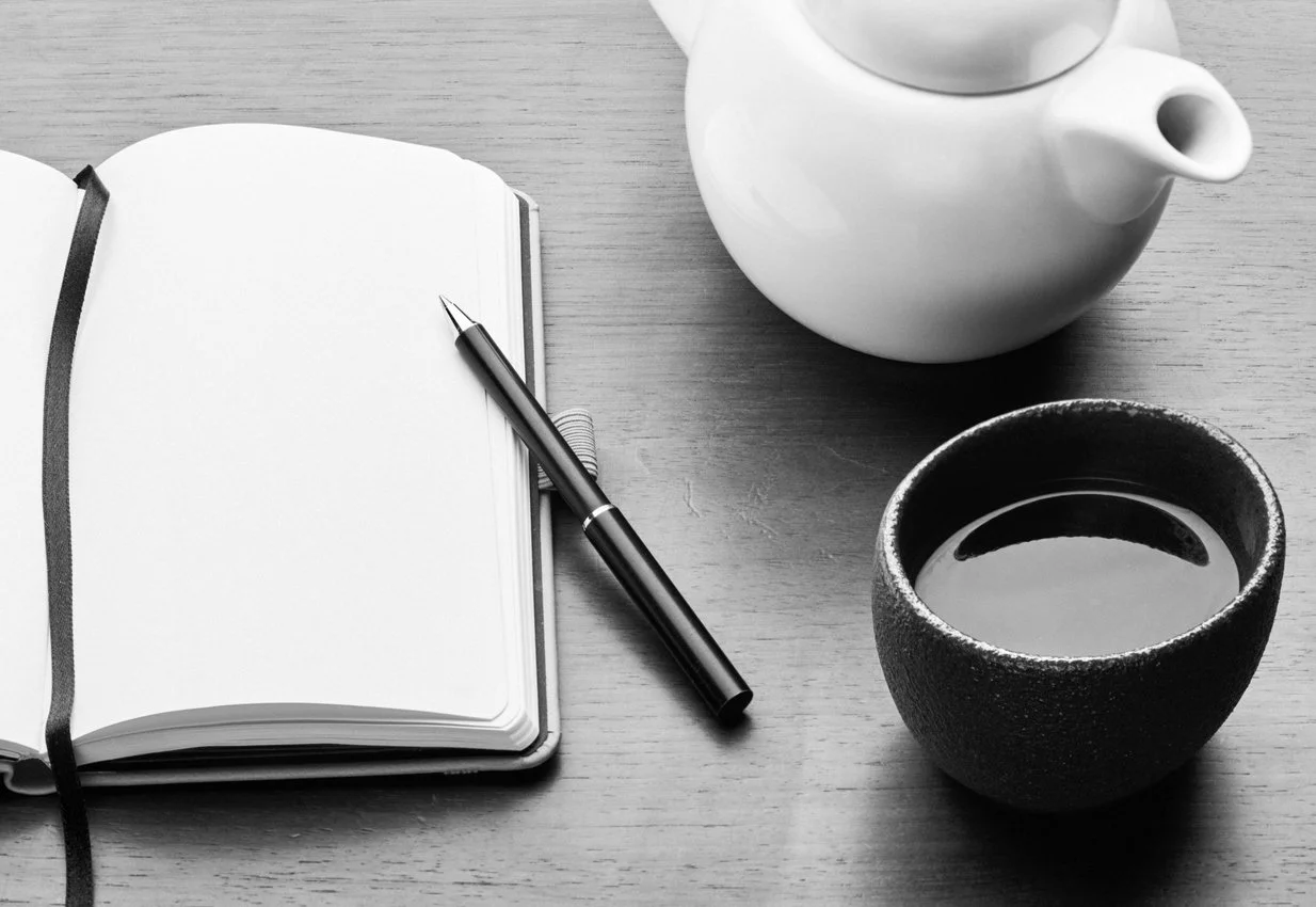 A black and white photo of an open blank notebook with a pen on top, a white teapot, and a cup of coffee on a wooden surface.