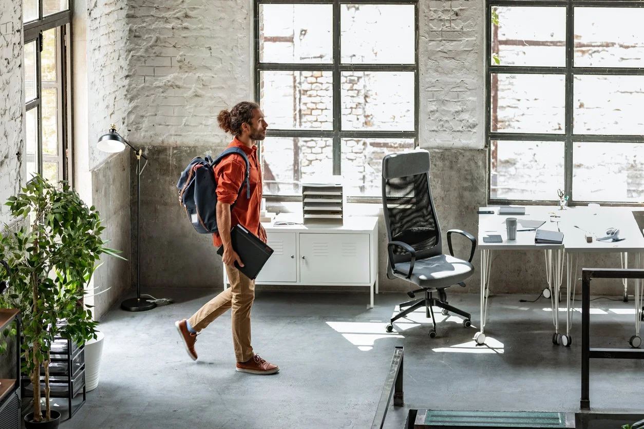 Young man with a backpack and a laptop entering a modern office space with large windows, an office chair, a desk with a coffee cup, and a potted plant.