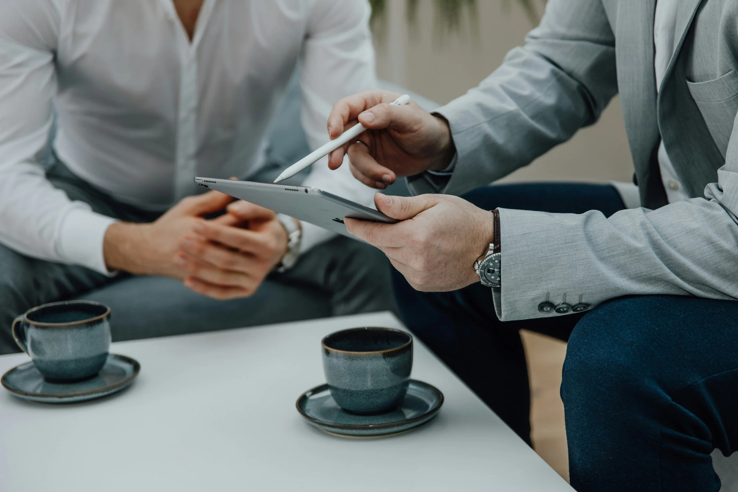 Two men in business attire having a discussion, one holding a tablet and a stylus while the other holds a cup of coffee.
