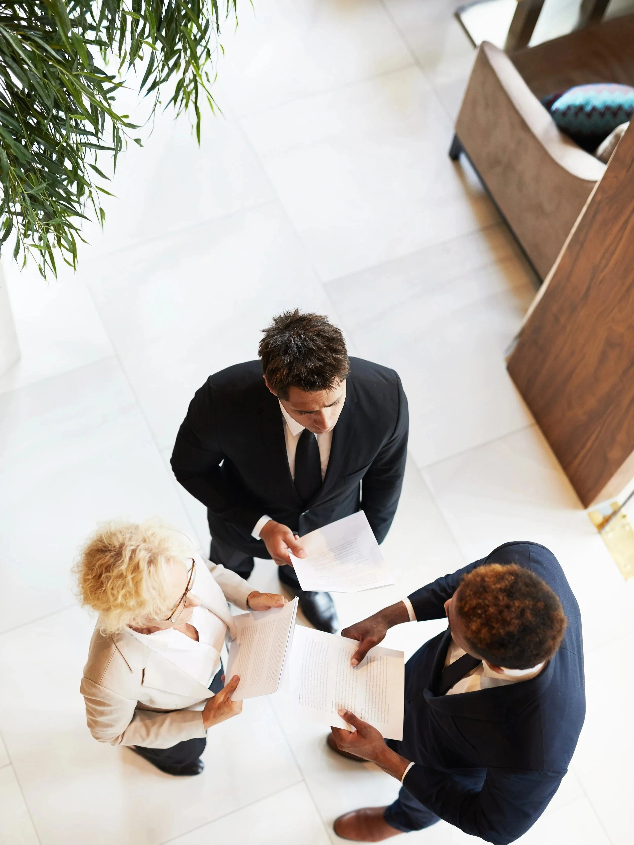 Three business professionals, two men and one woman, are standing in a lobby or reception area, engaged in a discussion, holding papers.