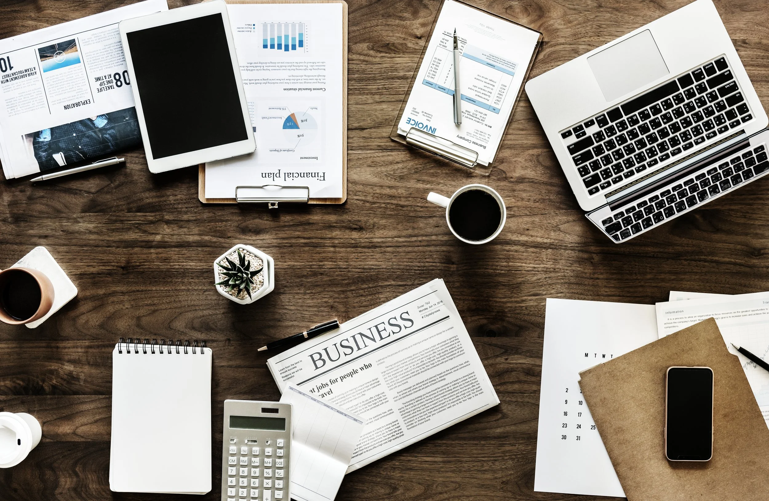 Overhead view of a cluttered wooden desk with laptop, tablets, coffee cups, business newspaper, notepad, calculator, documents, and a small potted plant.