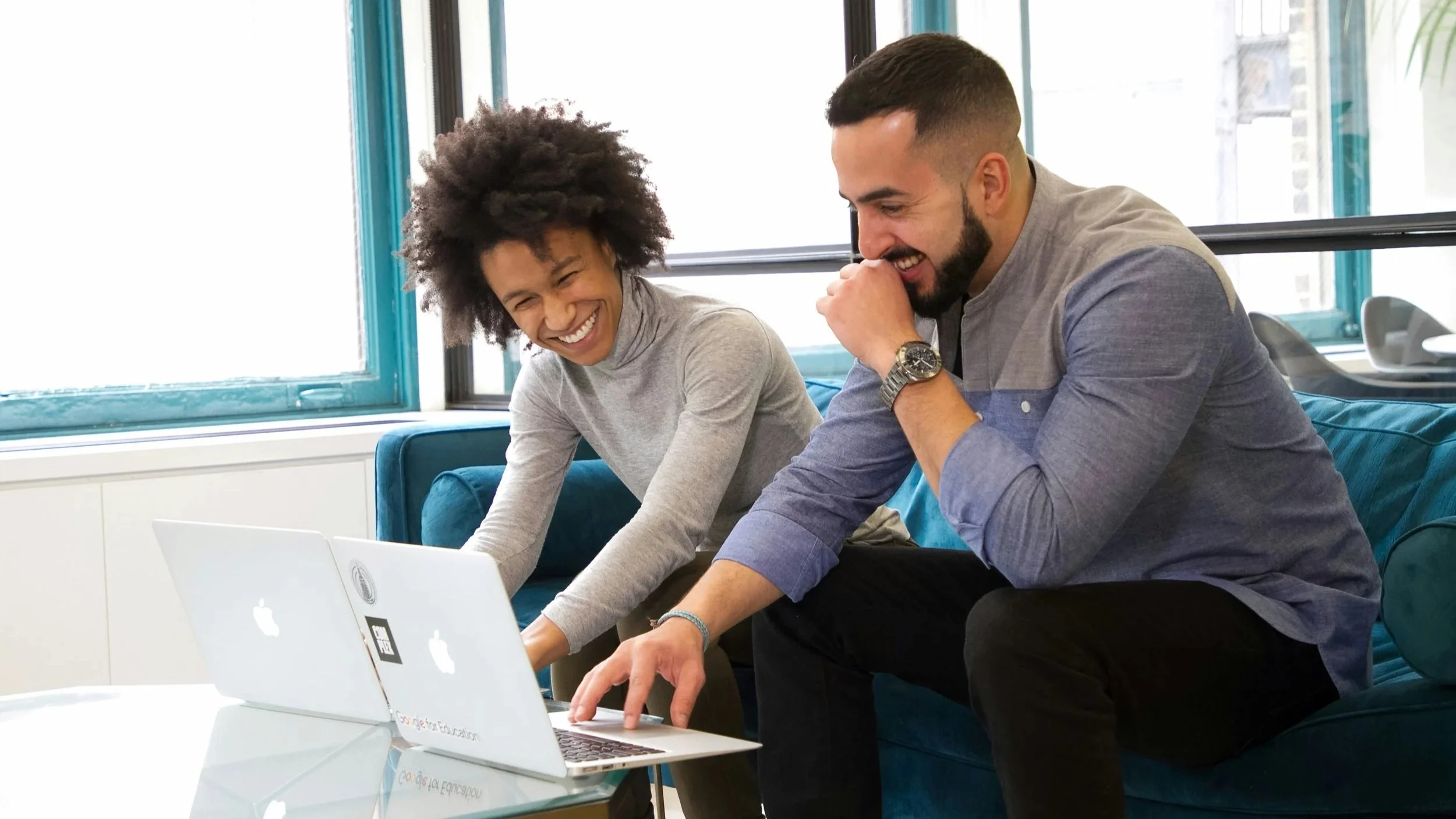 Two people sitting on a couch, laughing and looking at a laptop, in a bright room with large windows.