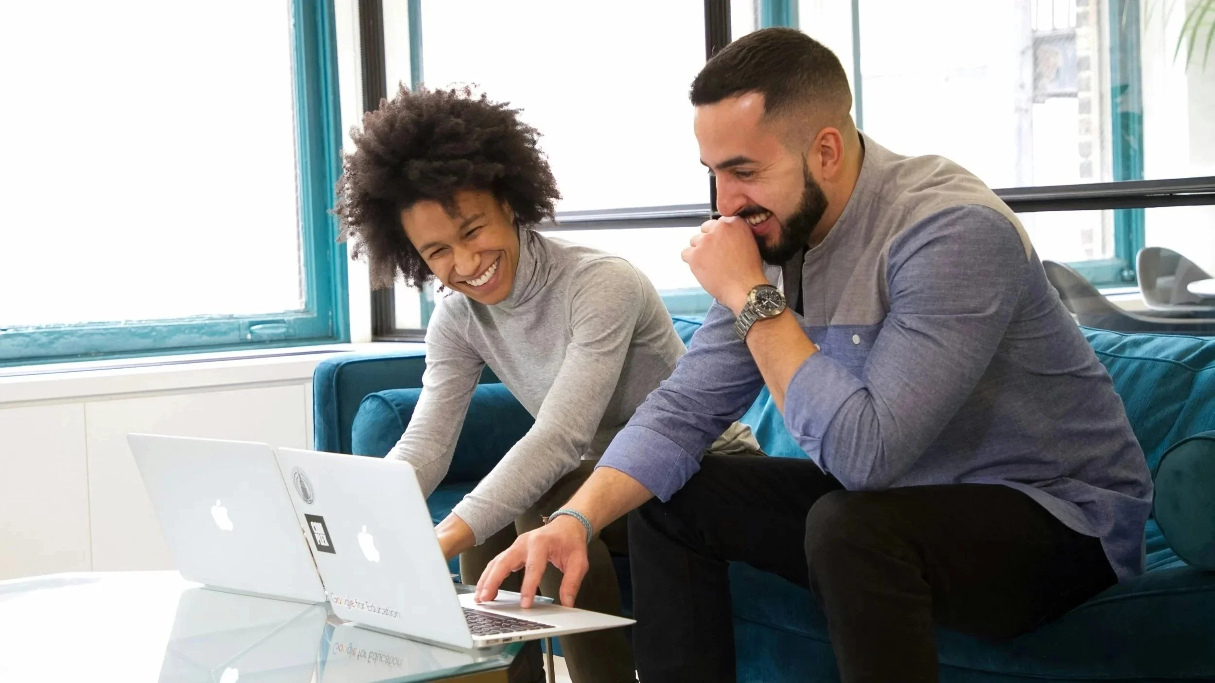 Two young adults sitting on a blue couch, laughing and working together on two laptops, in a bright office space with large windows.