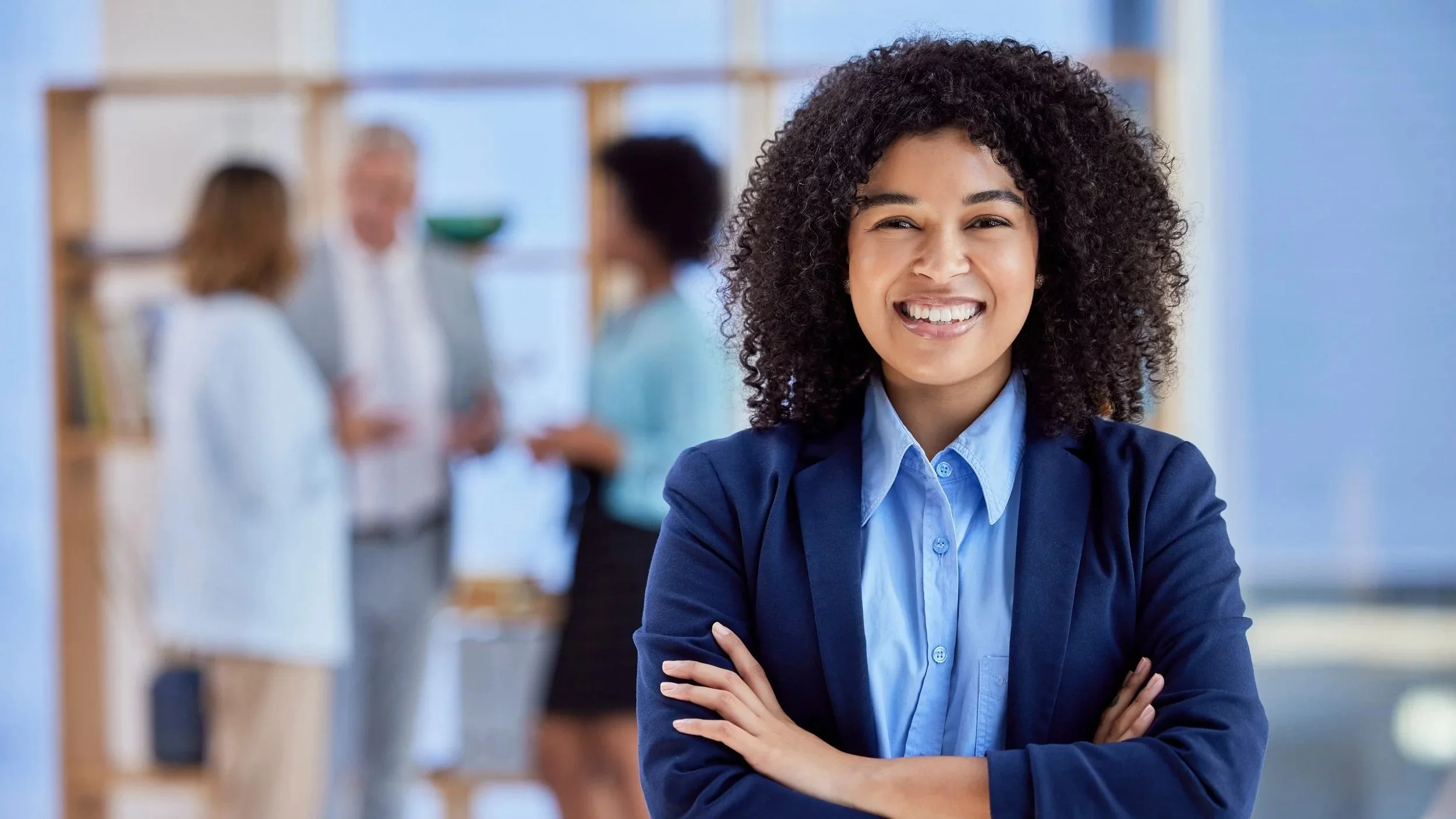 A woman with curly hair smiling and crossing her arms in an office setting with three blurred people in the background