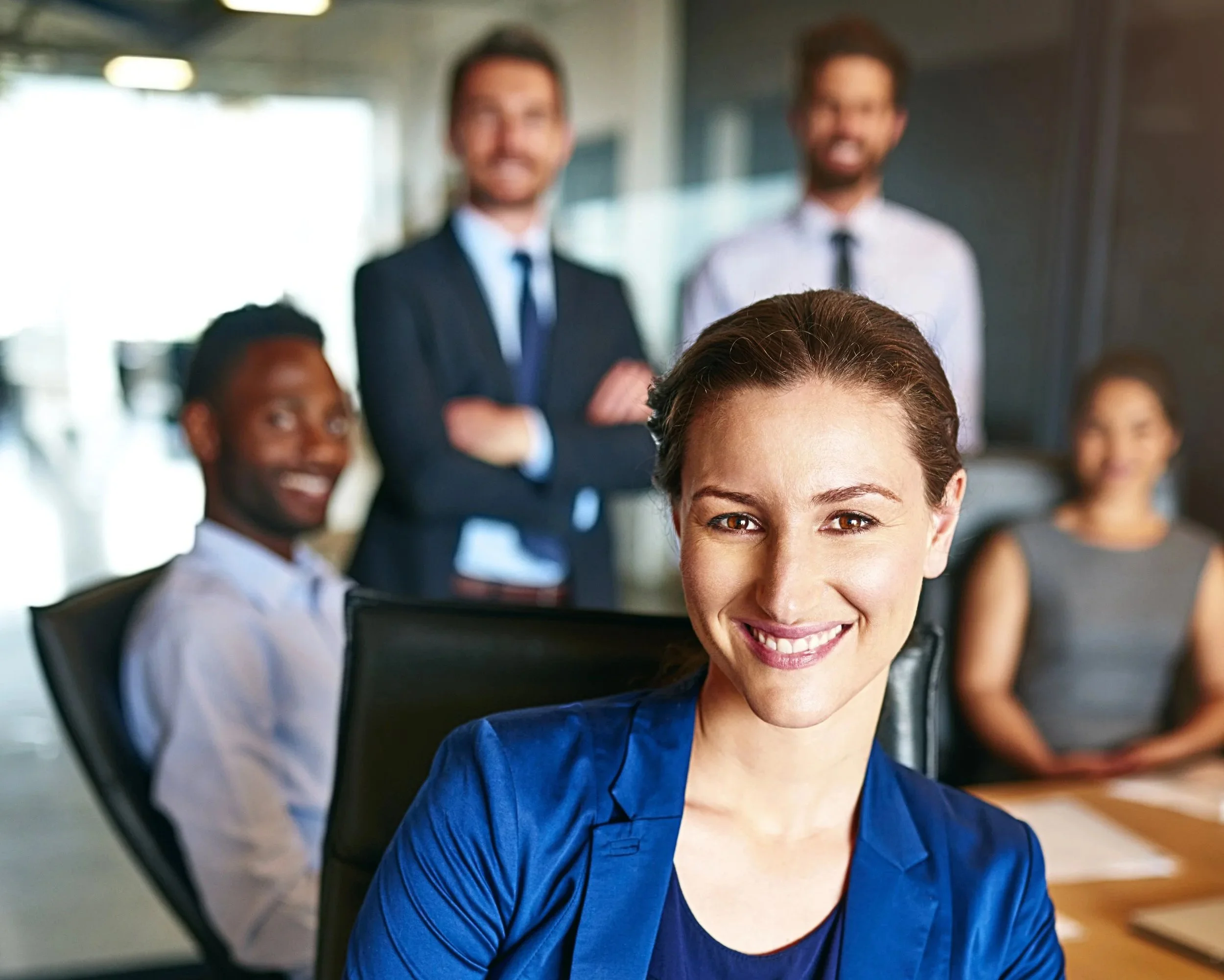A group of five diverse business professionals in a modern office, with one woman in the foreground smiling at the camera and four others in the background, some sitting and some standing, dressed in business attire.