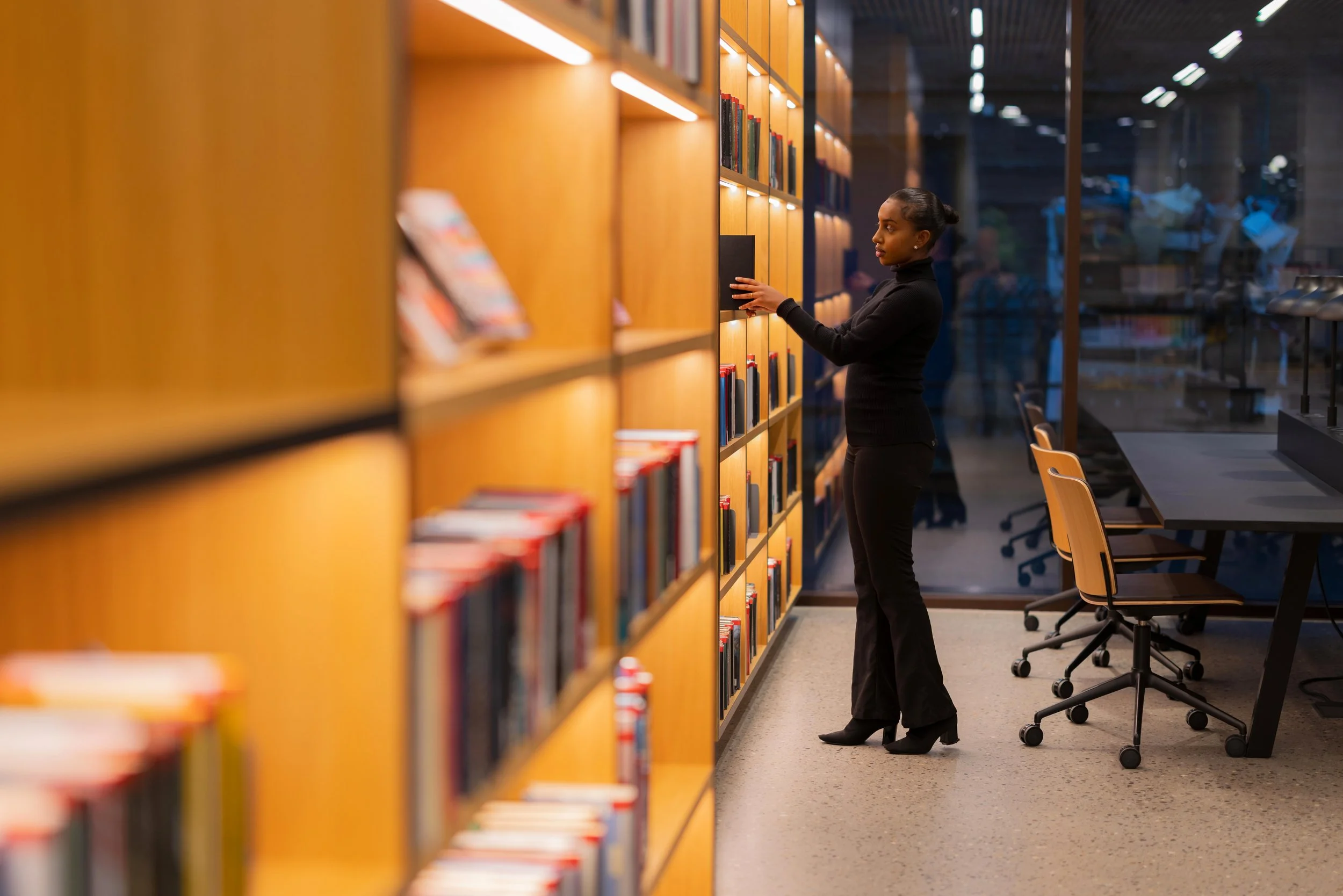 Woman browsing books in a library, standing beside a wooden bookshelf, wearing black clothing, with a glass wall and empty chairs in the background.