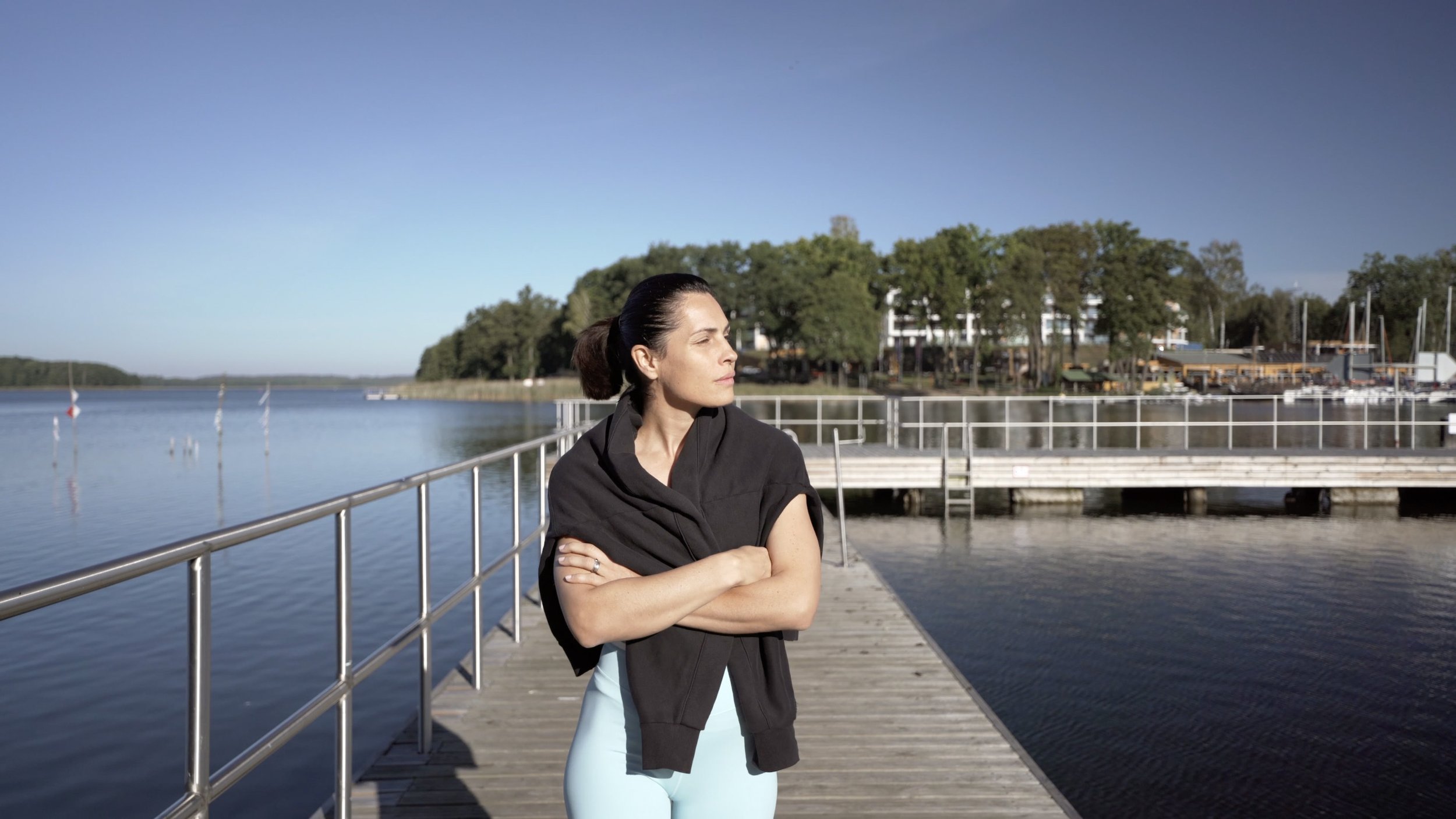 Woman standing on a wooden dock by a calm river, with trees and buildings in the background, looking to the side with arms crossed.