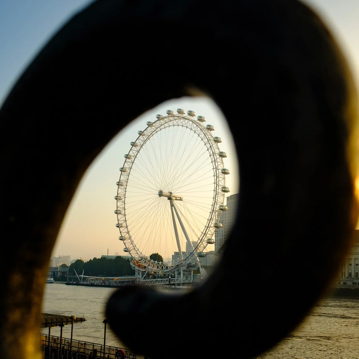 View of the London Eye through a handrail loop, with water and buildings in the background at sunset.