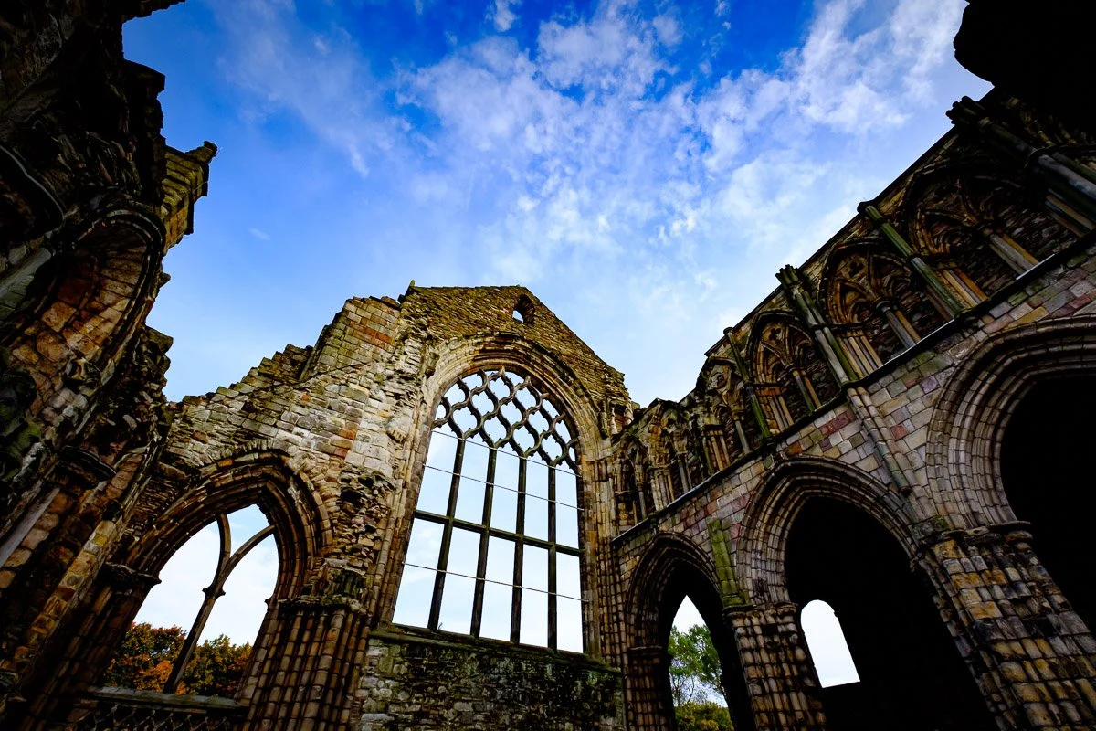View of a historic, Gothic-style stone church ruin with large arched windows and detailed stonework under a partly cloudy sky.