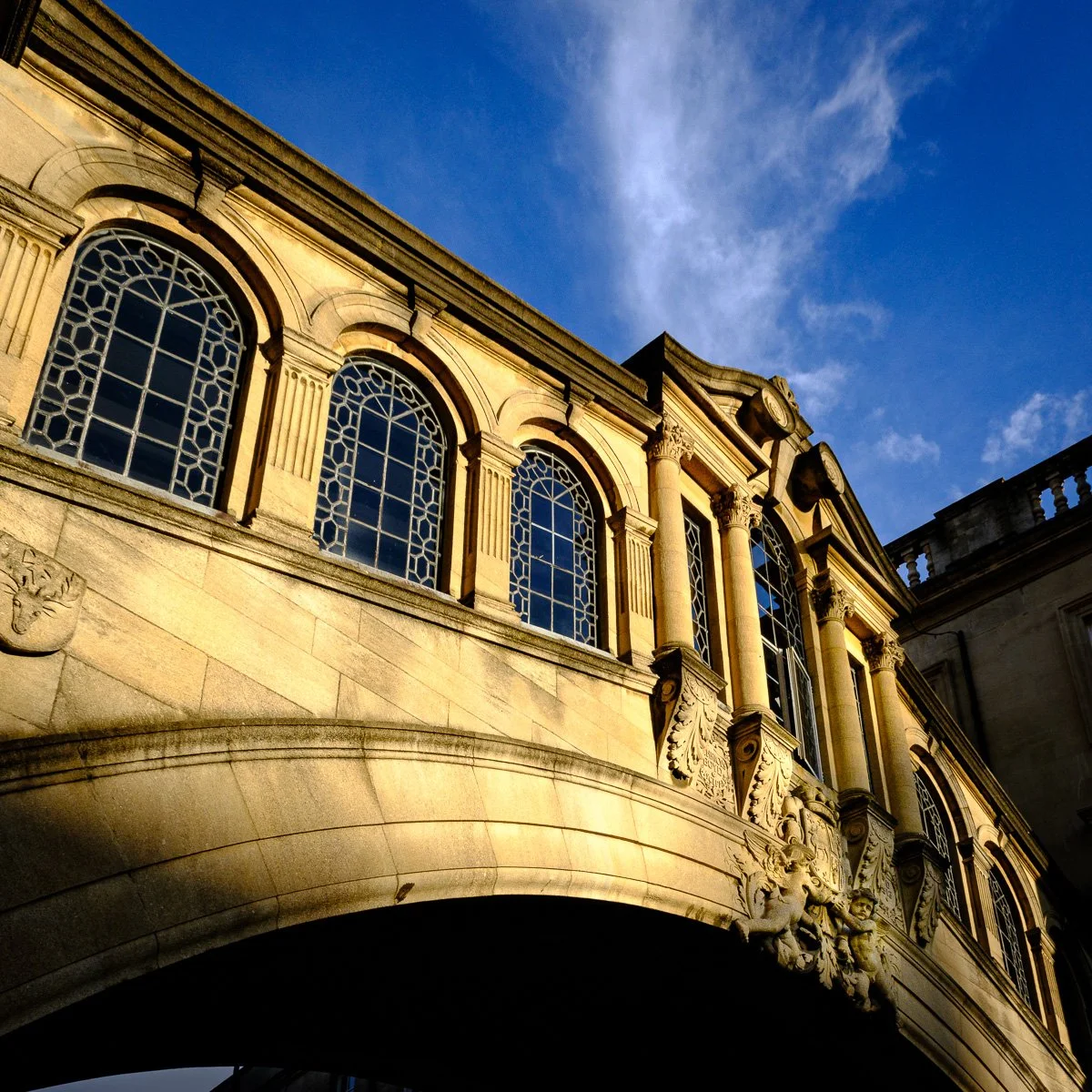 Elegant historic "Bridge of Sighs" in oxford with large arched windows and decorative stone carvings, bathed in warm sunlight under a blue sky with wispy clouds.