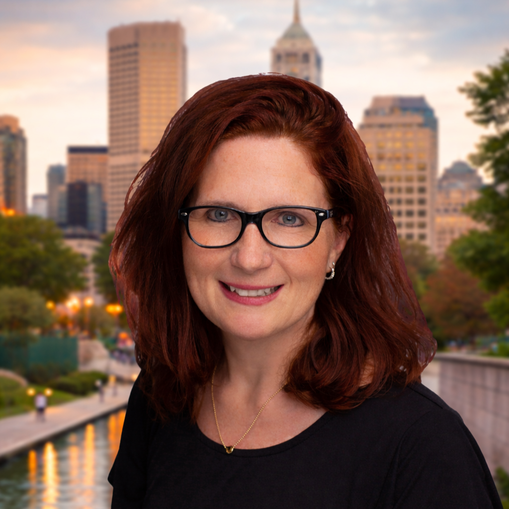 A woman with red hair, glasses, and a black top standing in front of a city skyline and a river at sunset.