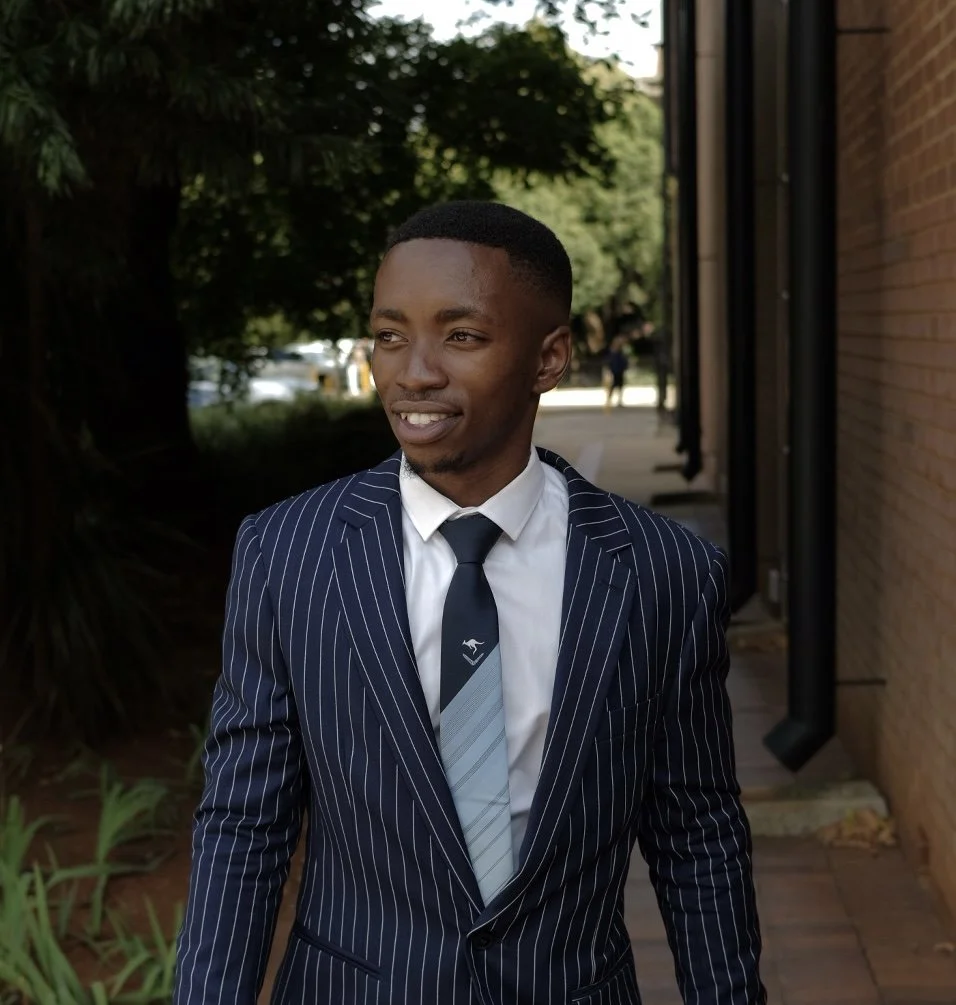 A young man in a navy blue pinstripe suit, white shirt, and striped tie walking outside near a brick building, with trees and sidewalk in the background.