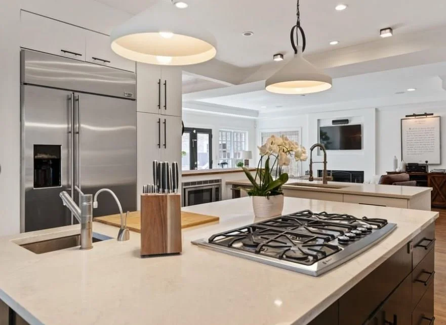 Modern kitchen with white countertops, stainless steel refrigerator, wooden knife block, orchid plant on the counter, and stove with gas burners. Open layout with living room visible in the background.
