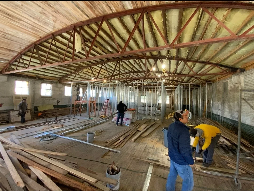 Interior view of a building under construction, with exposed wooden roof and metal framing, workers installing wood and metal components, and construction tools and materials scattered on the floor.