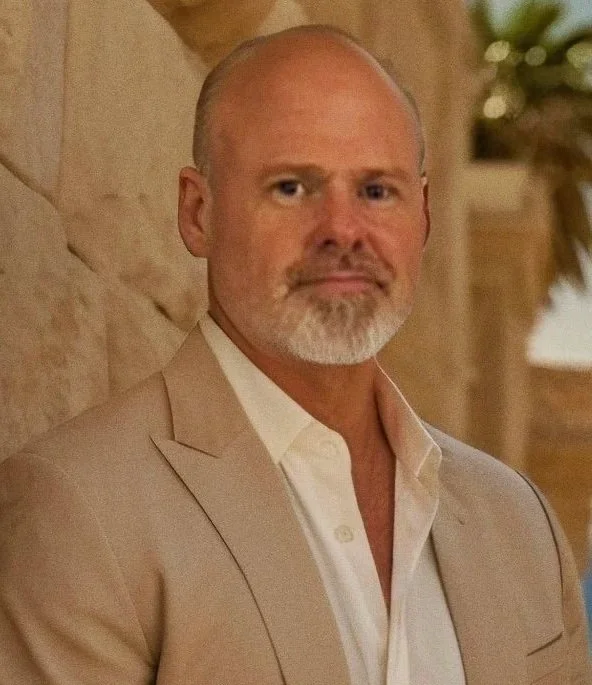 A middle-aged man with a beard and mustache, dressed in a beige suit and white shirt, standing indoors against a stone wall.