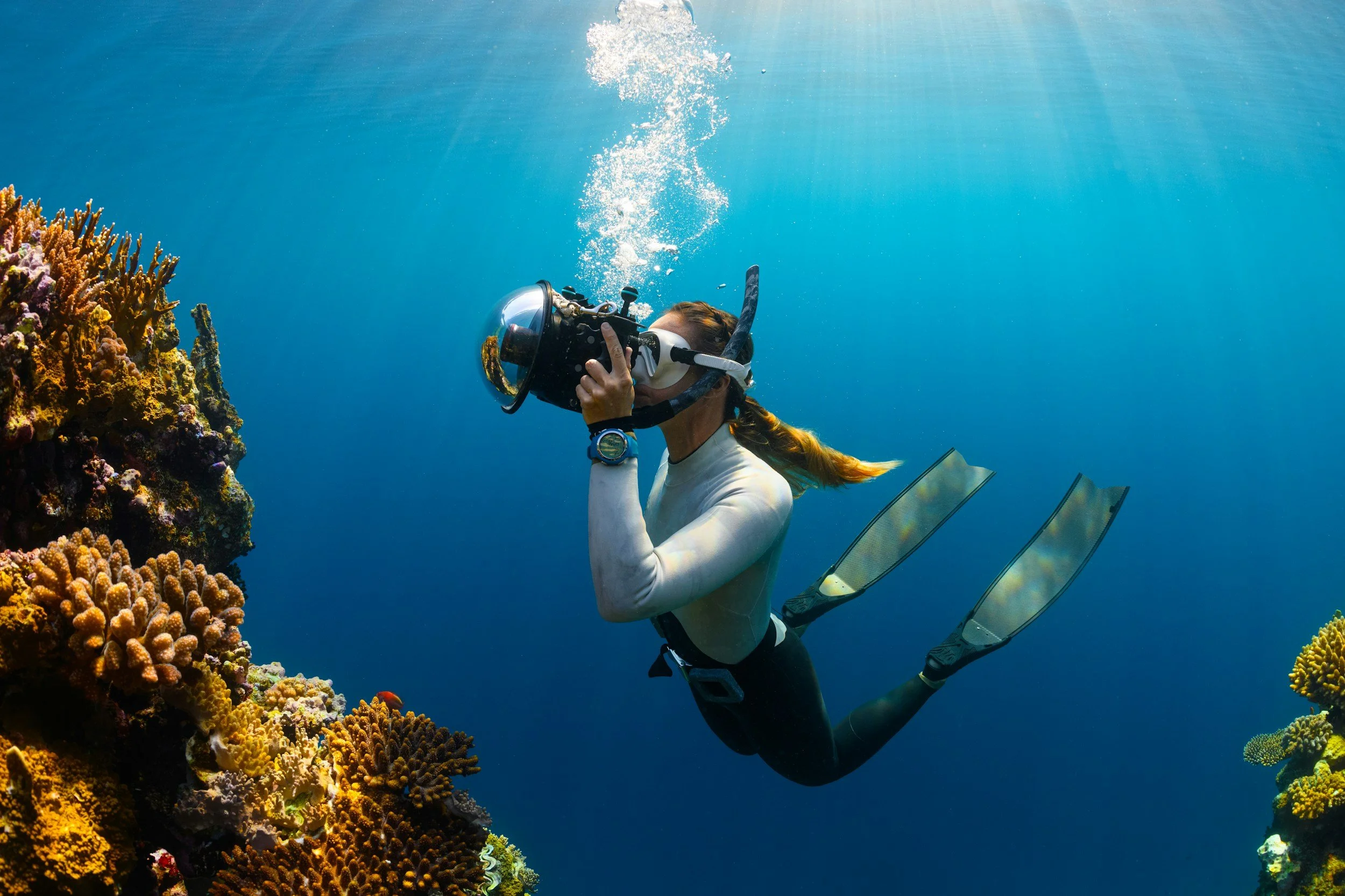 A woman scuba diving near coral reefs, holding a camera to take underwater photos, with sunlight shining through the water.