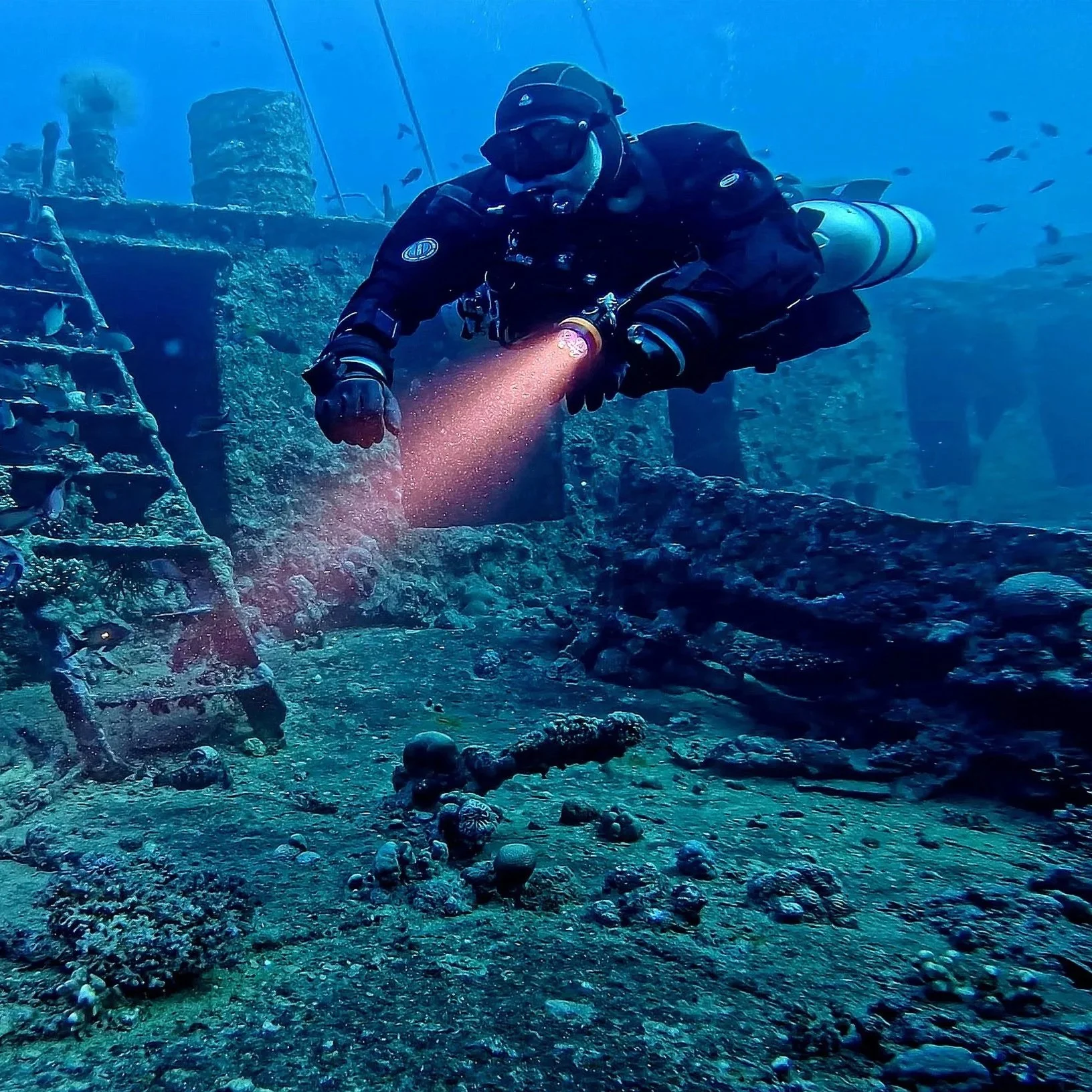 Taucher erkundet ein versunkenes Schiff unter Wasser, umgeben von kleinen Fischen und Meeresboden mit Steinen und Muscheln.