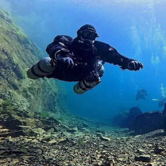 Taucher schwimmt im klaren Wasser über felsigem Grund, umgeben von blauer Wasserfarbe, mit einem weiteren Taucher im Hintergrund.