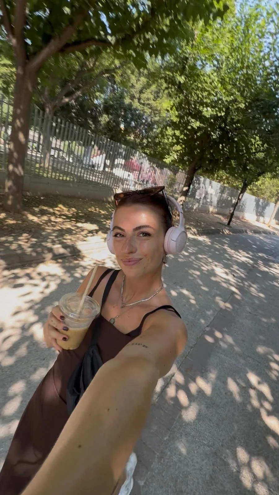 A young woman with dark hair, wearing large pink headphones, smiling and taking a selfie while walking outdoors on a sunny day. She is holding an iced coffee and is dressed in a dark sleeveless top with sunglasses on her head. She is under trees with shadows cast on the sidewalk, and a fence is visible in the background.