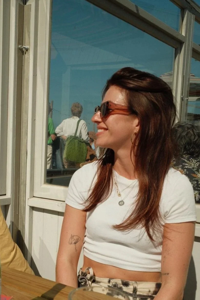A woman with long reddish-brown hair wearing sunglasses, a white crop top, and patterned pants, smiling while sitting at a table with a reflection of other people behind a glass window.