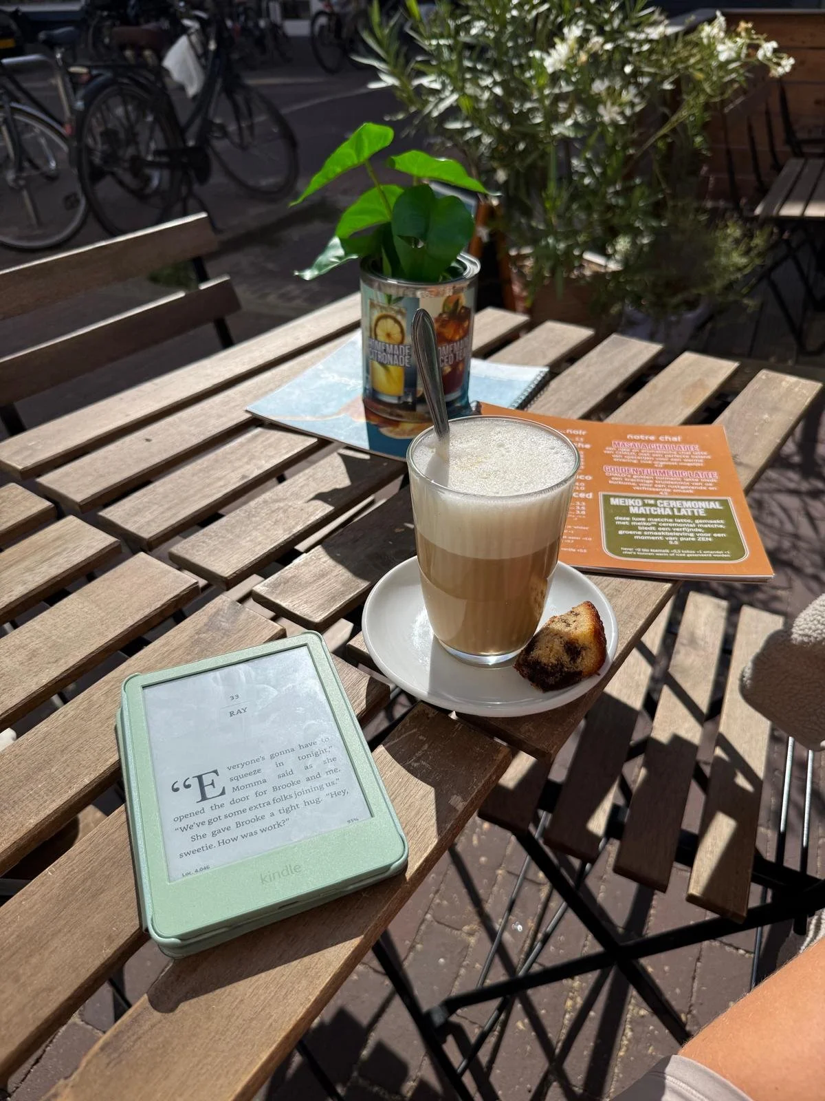 A wooden outdoor café table with a kindle e-book, a latte with foam, a slice of marble cake, a small potted plant, an orange flyer, and a menu. In the background, there are bicycles and outdoor seating.