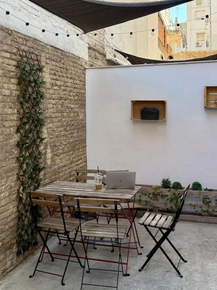 Outdoor patio with a wooden table and chairs, a laptop, a drink, a white wall, some plants, and string lights, with brick and white walls and umbrellas overhead.