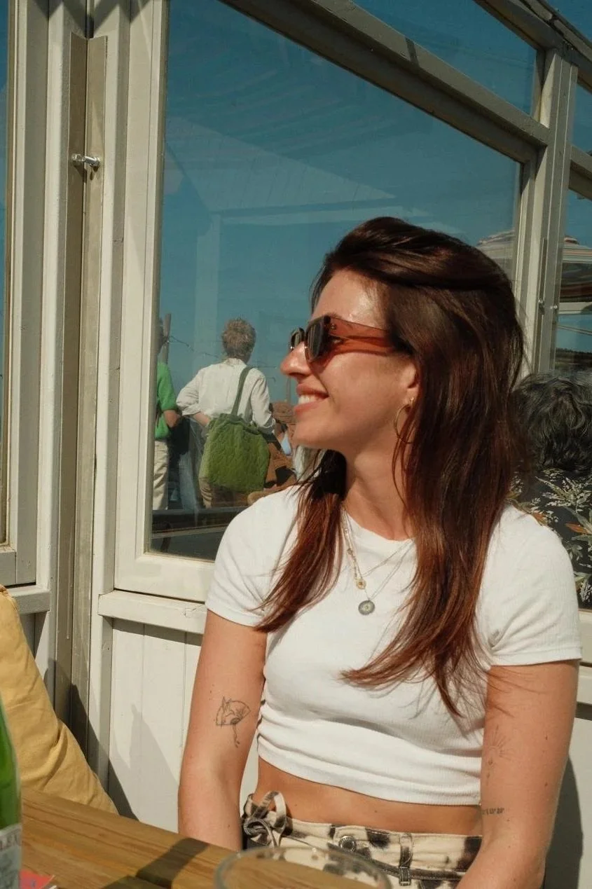 A woman with long brown hair, wearing sunglasses, a white crop top, and necklaces, sitting at a table outside near a glass window, smiling and looking to the side.