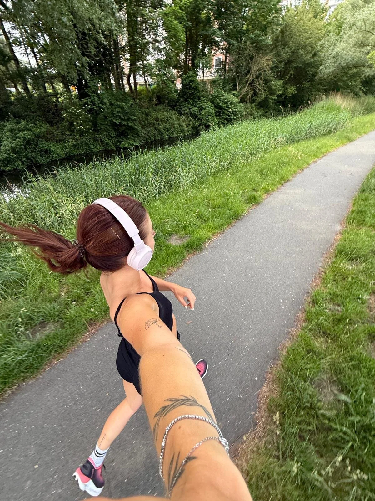 A woman jogging on a paved trail surrounded by green grass and trees, wearing headphones, a black tank top, and pink running shoes, with her arm extended toward the camera in a selfie-style photo.