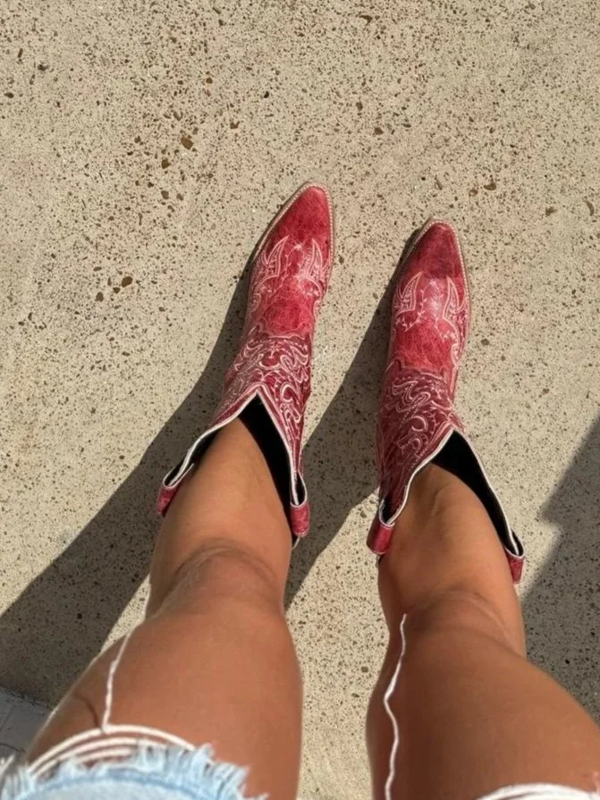 A person's legs wearing pink cowboy boots with white decorative stitching, standing on a textured beige concrete surface, with earbuds visible.