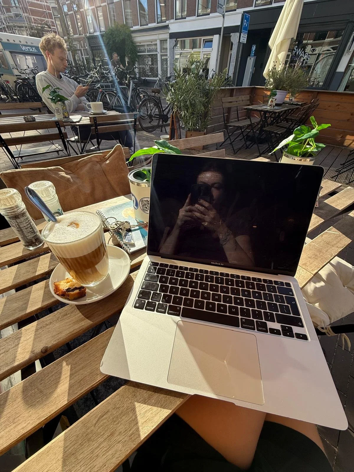 Outdoor cafe scene with a laptop, coffee, and a pastry on a wooden table, next to a person holding a phone. In the background, another person is sitting on a bench using a phone, with bicycles and buildings visible behind.