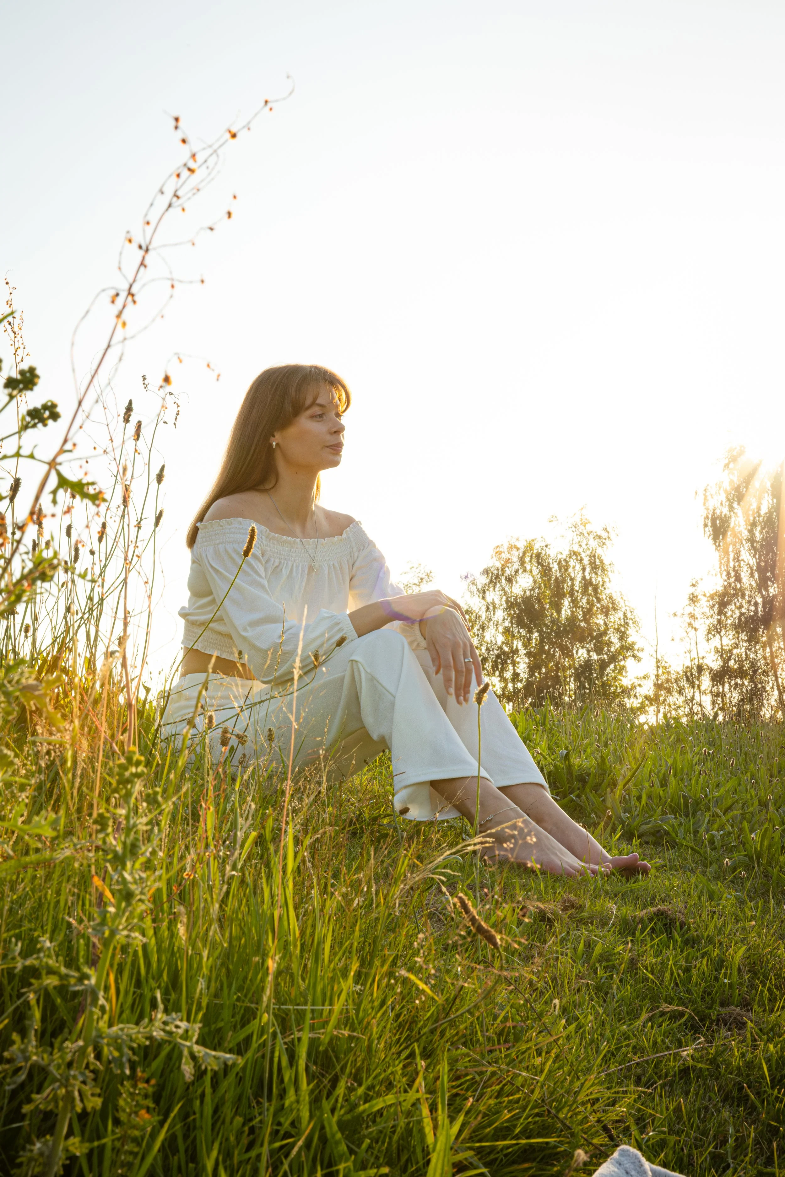 A young woman with long hair, wearing a white off-shoulder top and white pants, sitting barefoot on a grassy hill at sunset surrounded by wildflowers and trees.