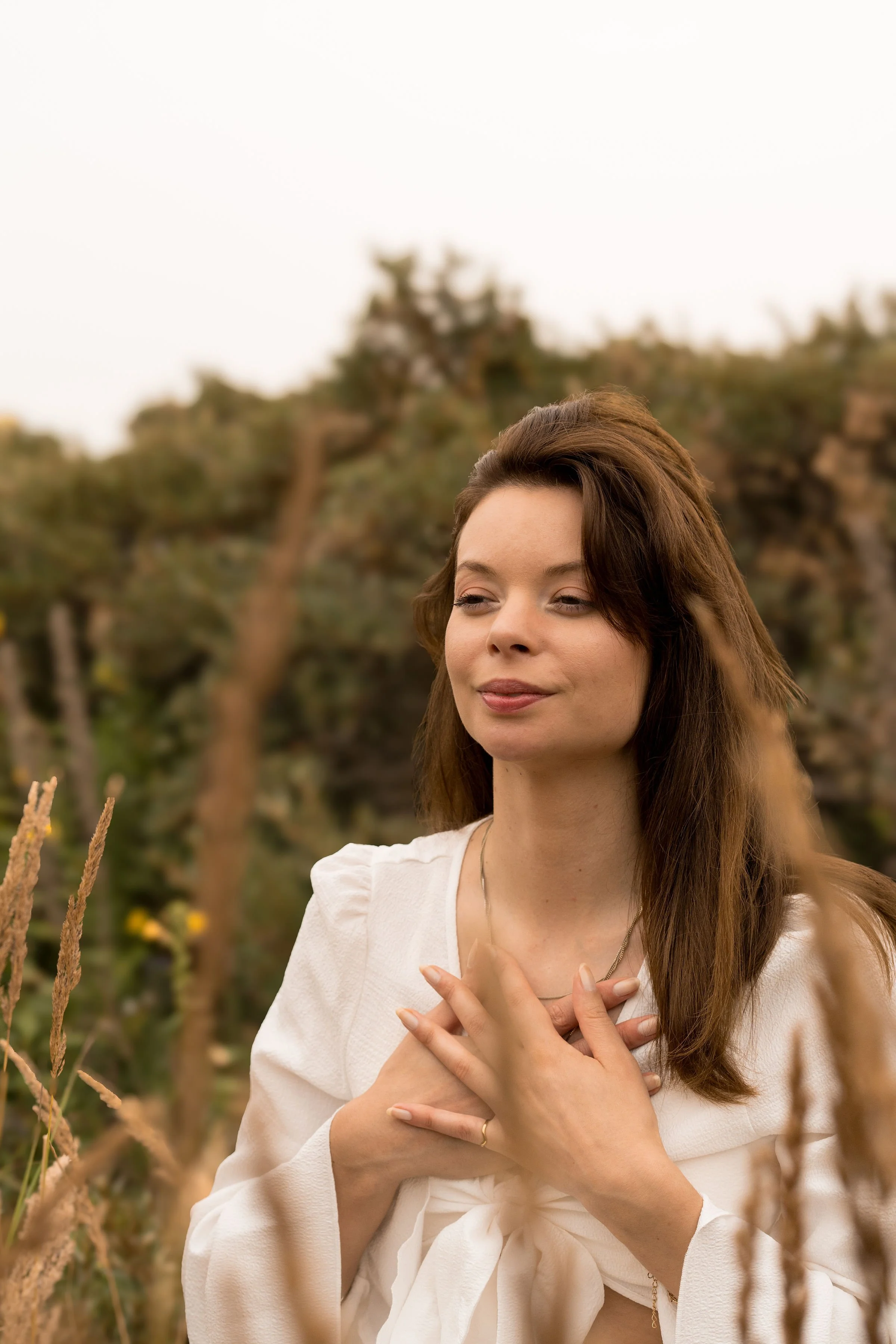 A woman with brown hair wearing a white top and a necklace, standing outdoors with her hands over her chest, surrounded by tall grass and trees in the background.