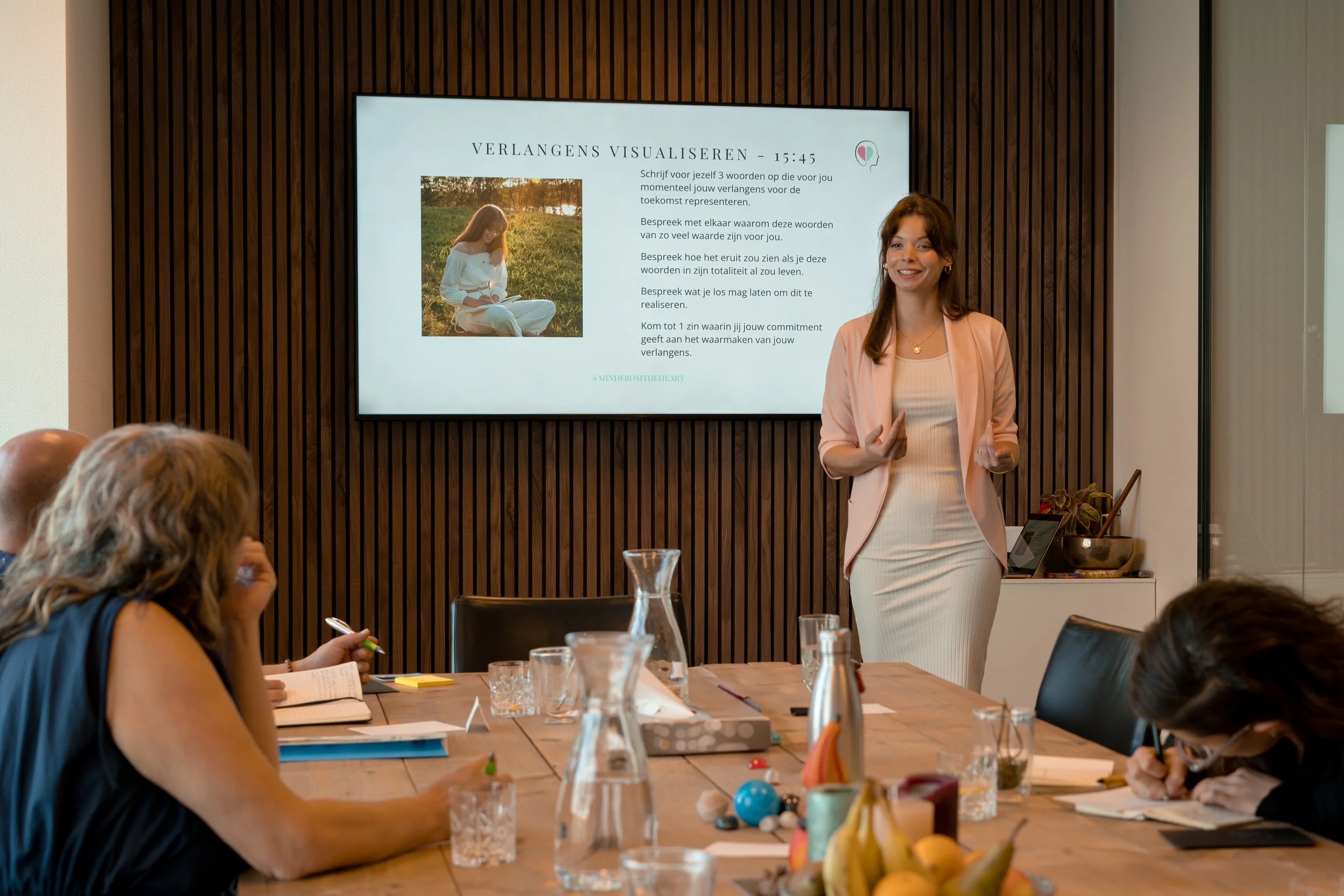 A woman giving a presentation in a conference room with a large screen on the wall behind her. Several attendees are seated at a table, taking notes and listening.