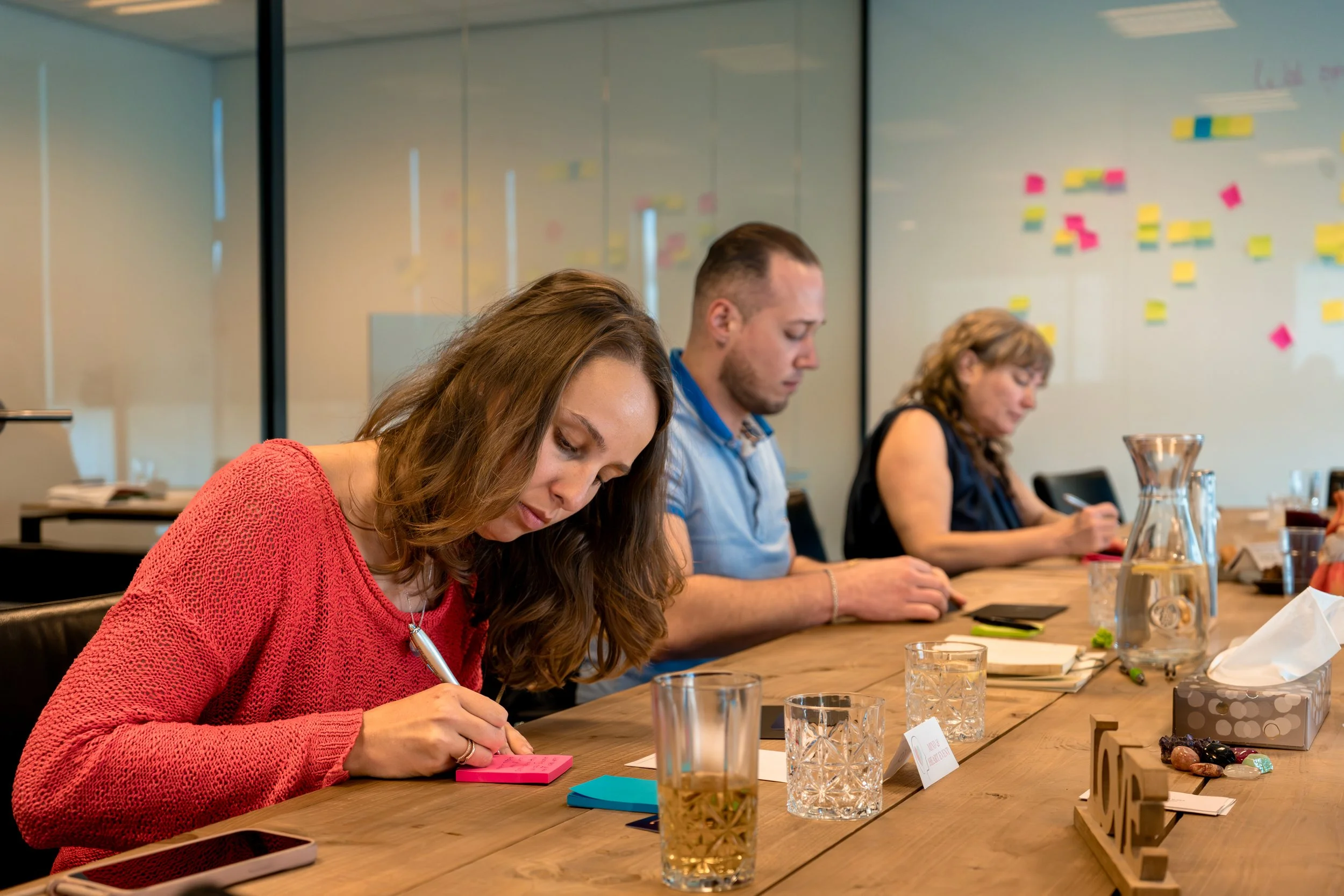 People sitting at a conference table, taking notes, in a meeting room with a whiteboard with sticky notes in the background.