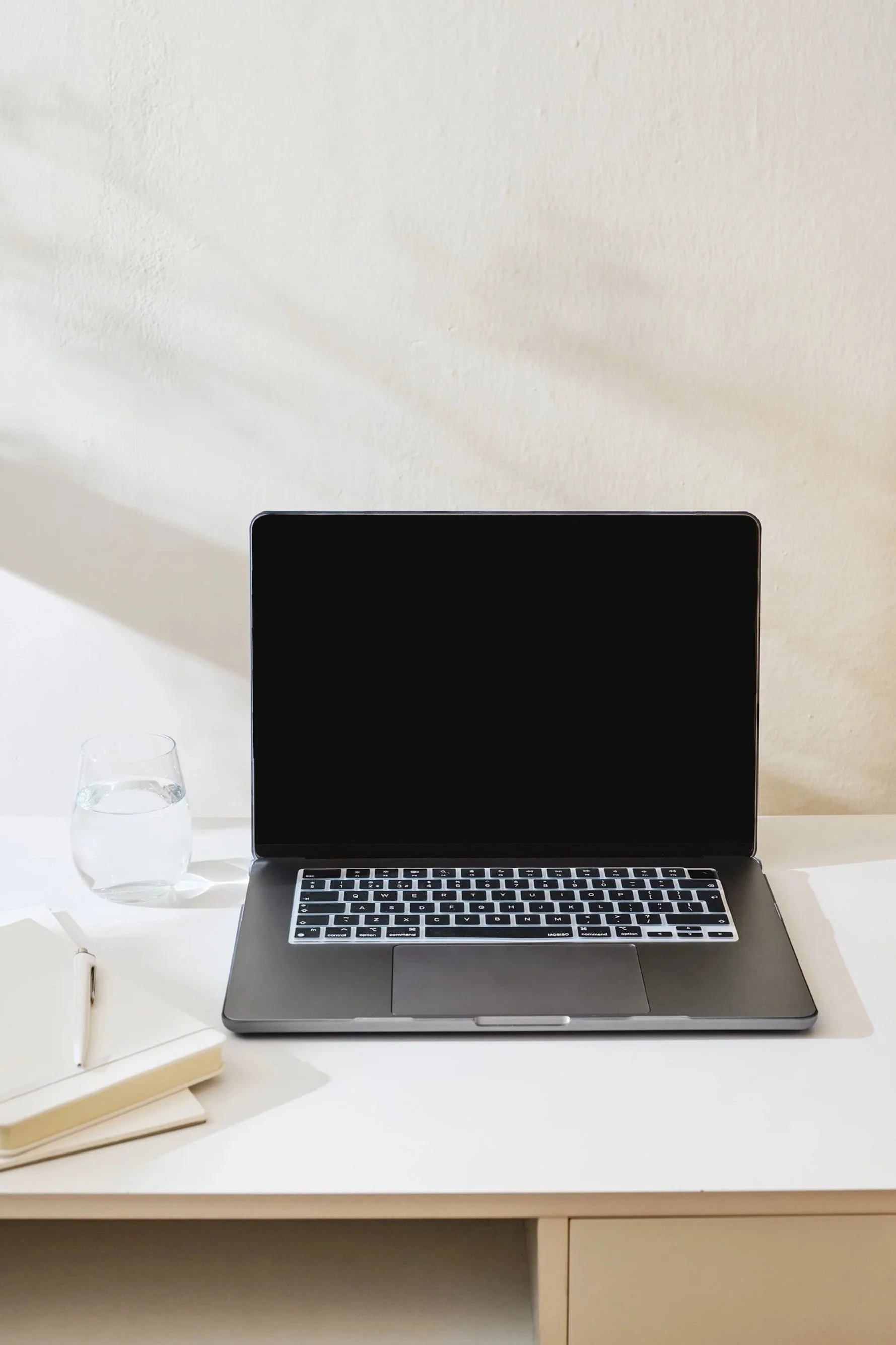 An open laptop, a glass of water, a white journal, and a white pen sit on top of a white desk. The desk sits in front of a tan wall, and light from the left side casts shadows on the wall and desk.